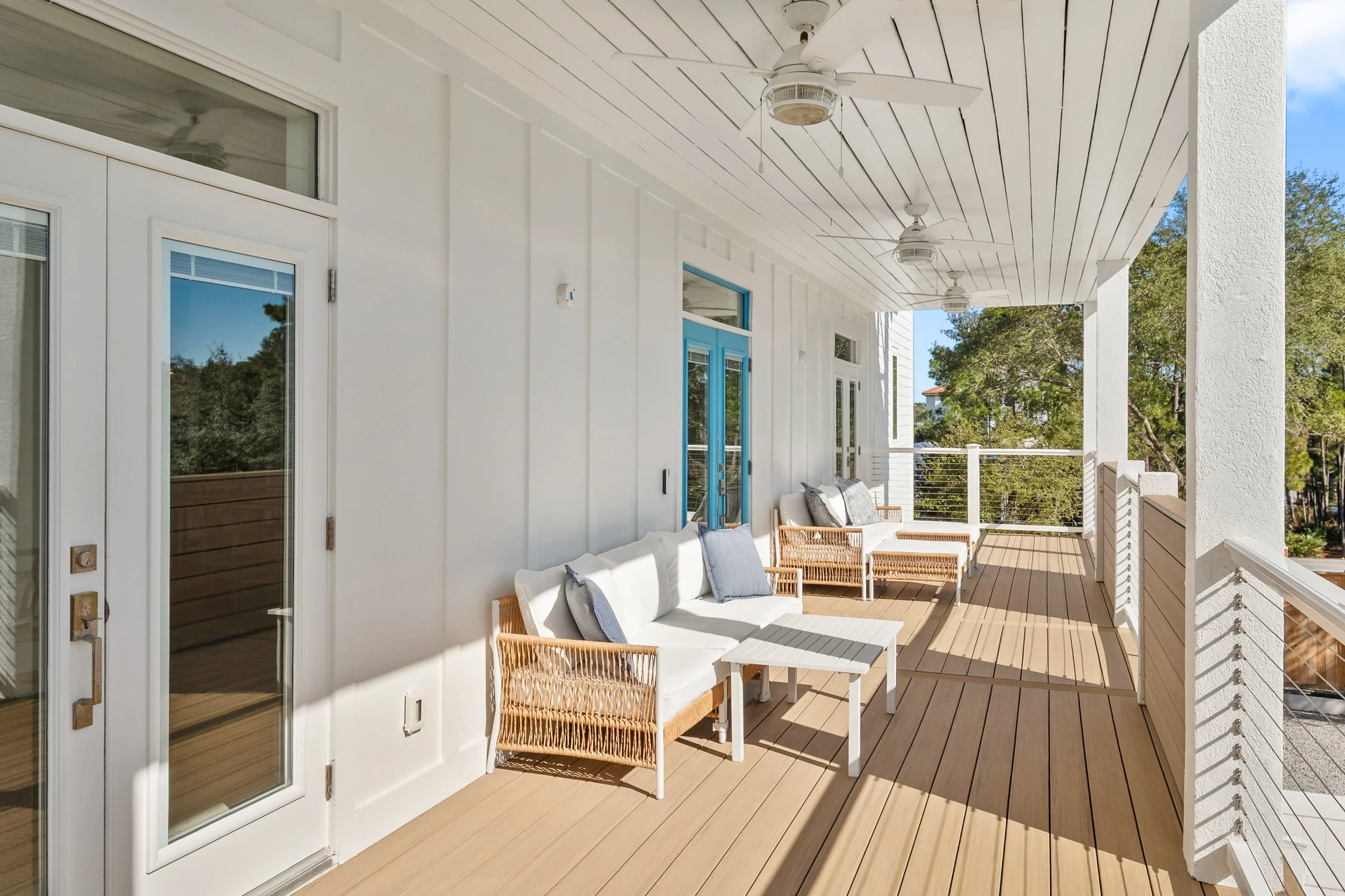 A spacious balcony with white walls and ceiling, featuring ceiling fans, beige wooden flooring, and outdoor furniture including white cushioned chairs with wicker arms, a small white table. The balcony is surrounded by a white railing, with trees vis