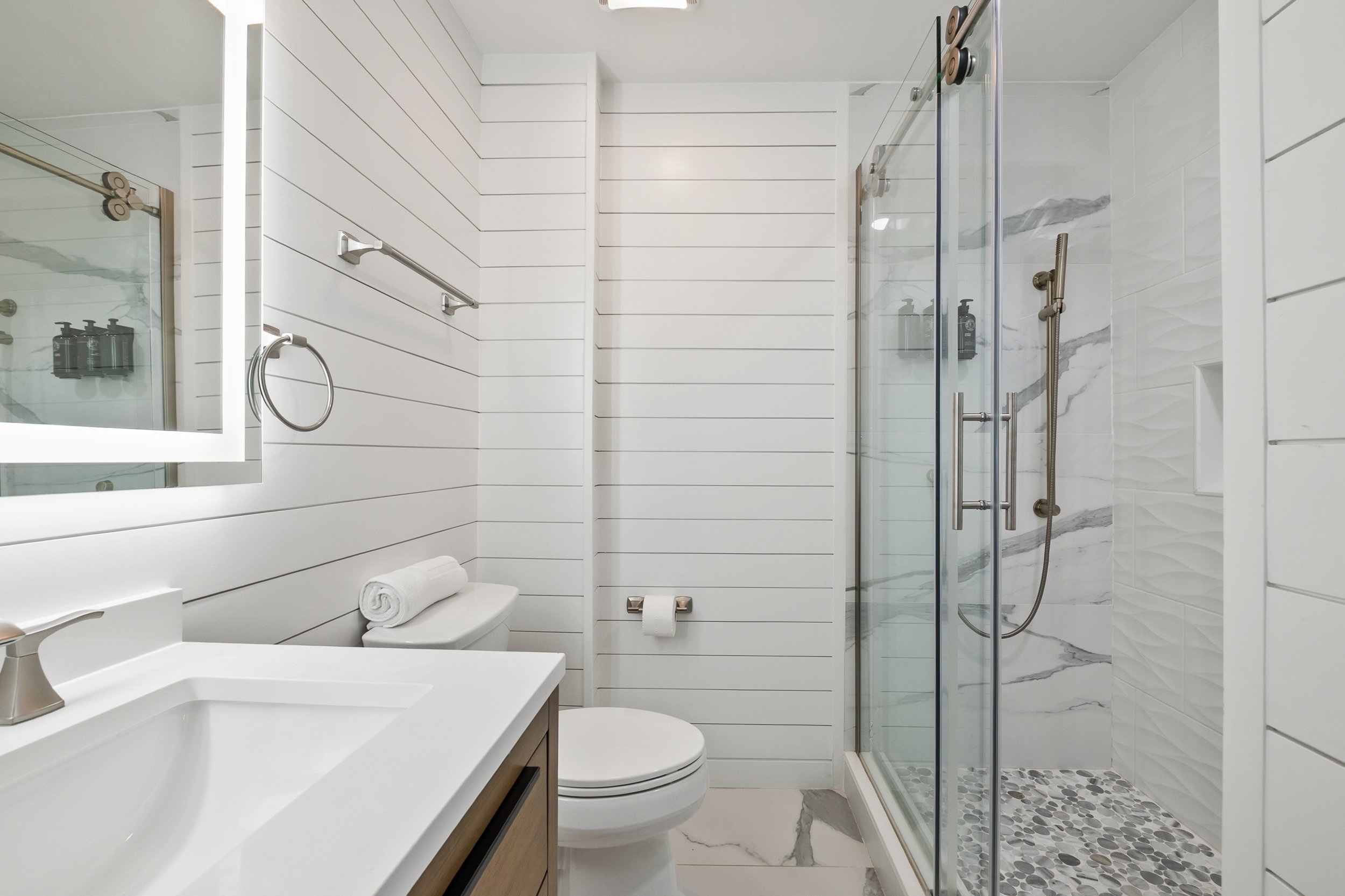 Modern bathroom with white shiplap walls, a white vanity with a sink, a wall mirror, a towel ring, a toilet with a rolled towel on top, and a glass-enclosed shower with marble tiles and pebble floor.