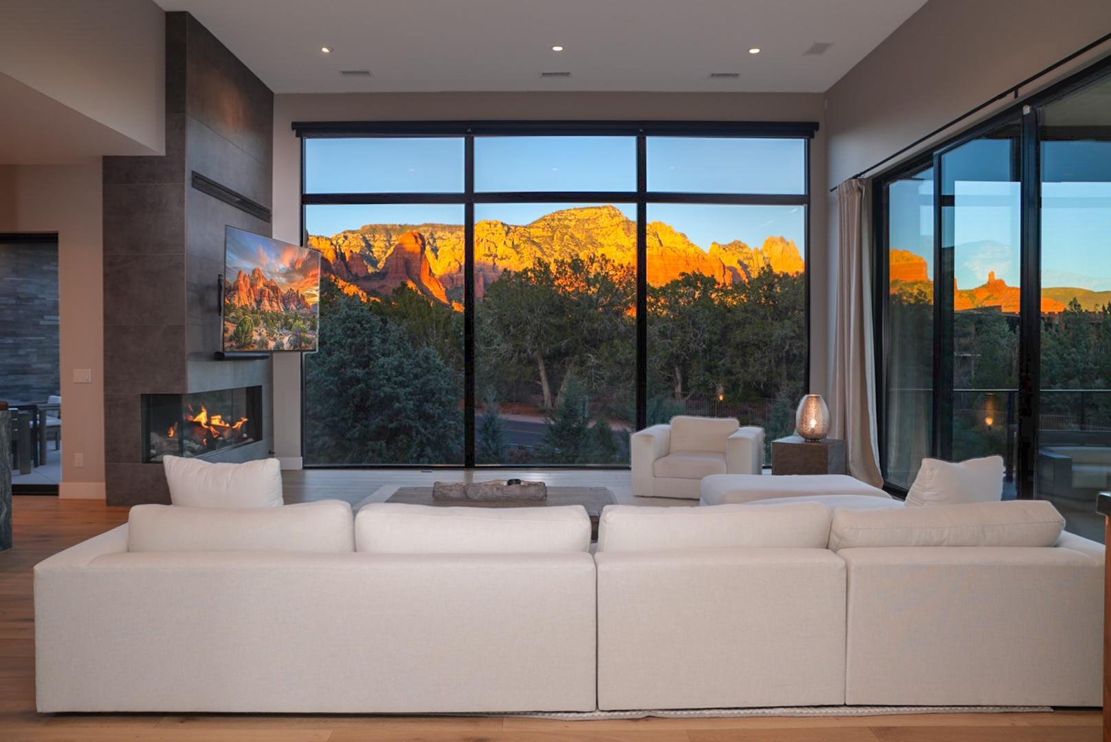 Modern living room with large glass windows showing a mountain range at sunset, white sofa, armchairs, fireplace, and a TV mounted on the wall.