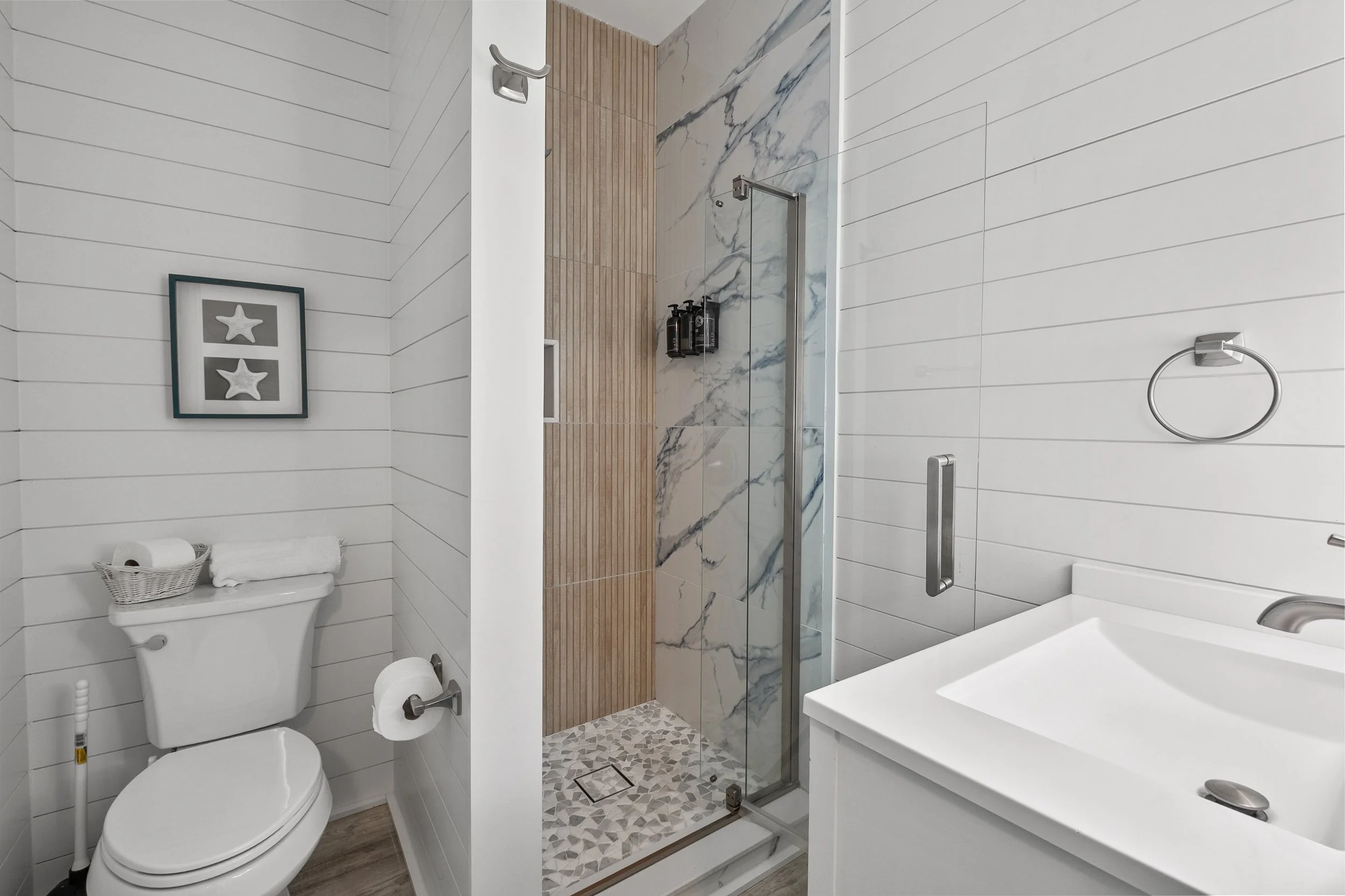 Modern bathroom featuring a white toilet, a glass-enclosed shower with marble and wooden accents, a white sink, and wall-mounted accessories.