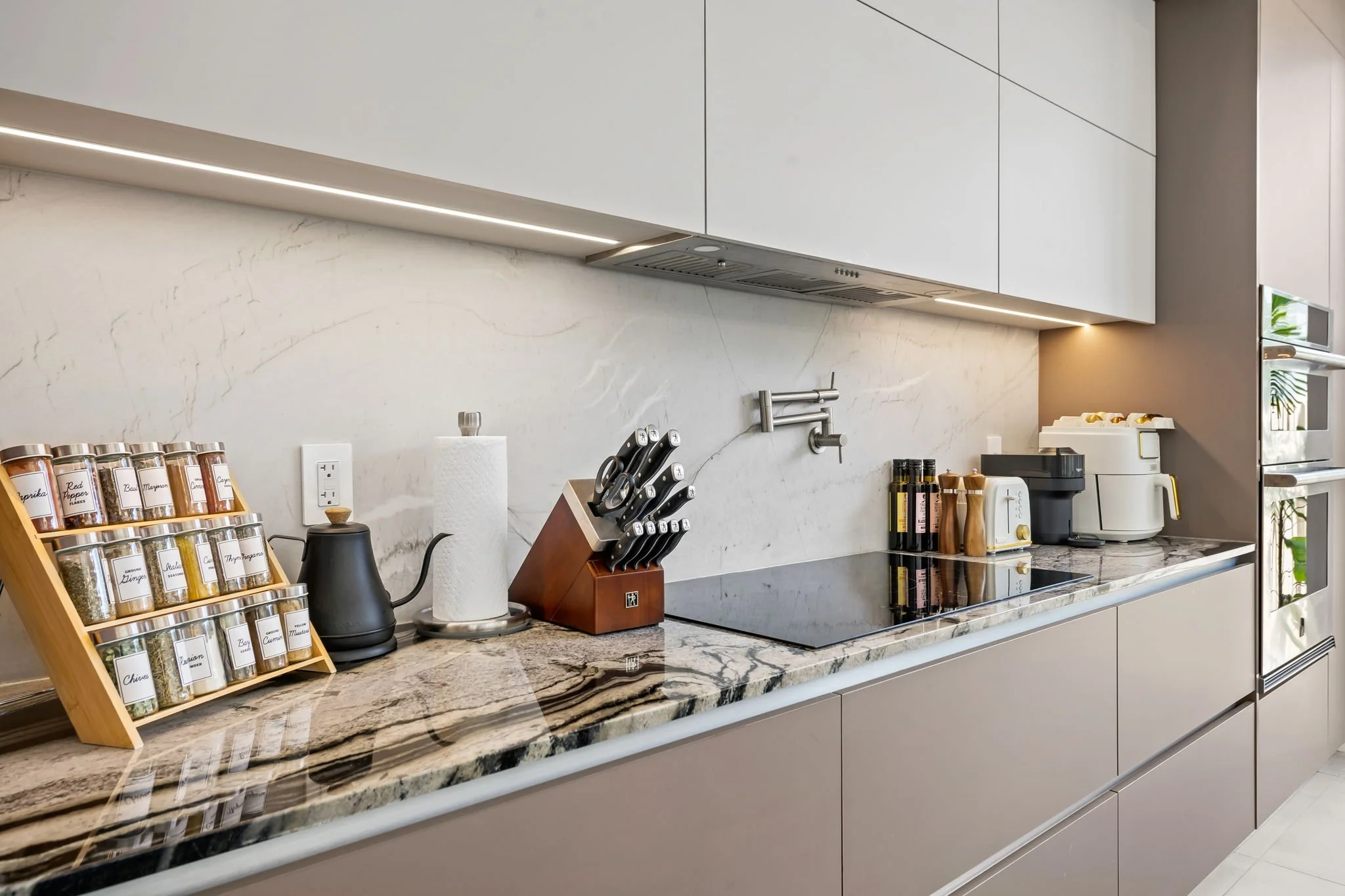 Kitchen countertop with spice jars, paper towel roll, knives in a wooden block, and small appliances including a toaster and coffee maker, with white cabinets and a marble backsplash.