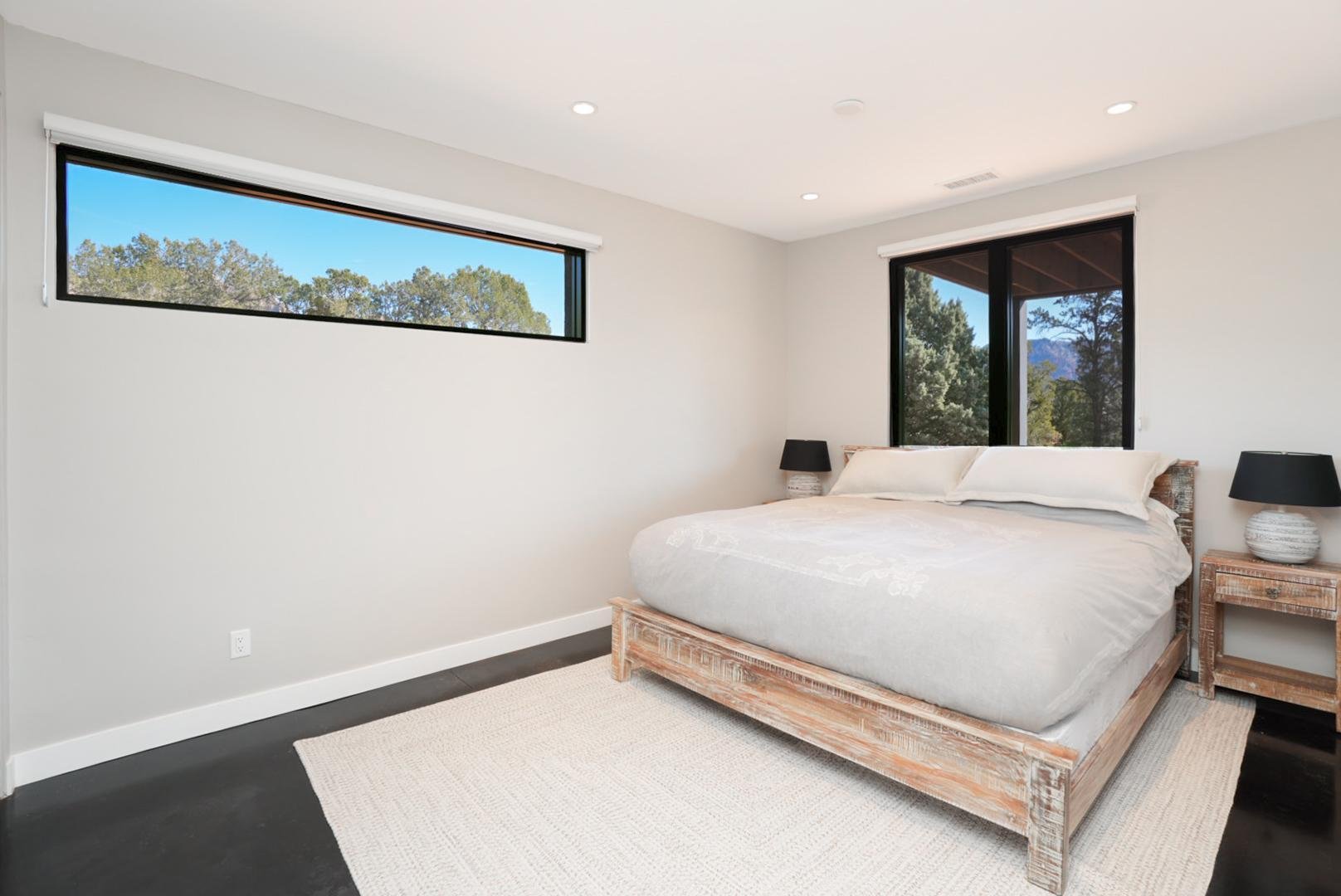 A bedroom with a wooden bed frame, white bedding, two black lamps on rustic wooden nightstands, a large window, and a smaller horizontal window showing trees and blue sky.