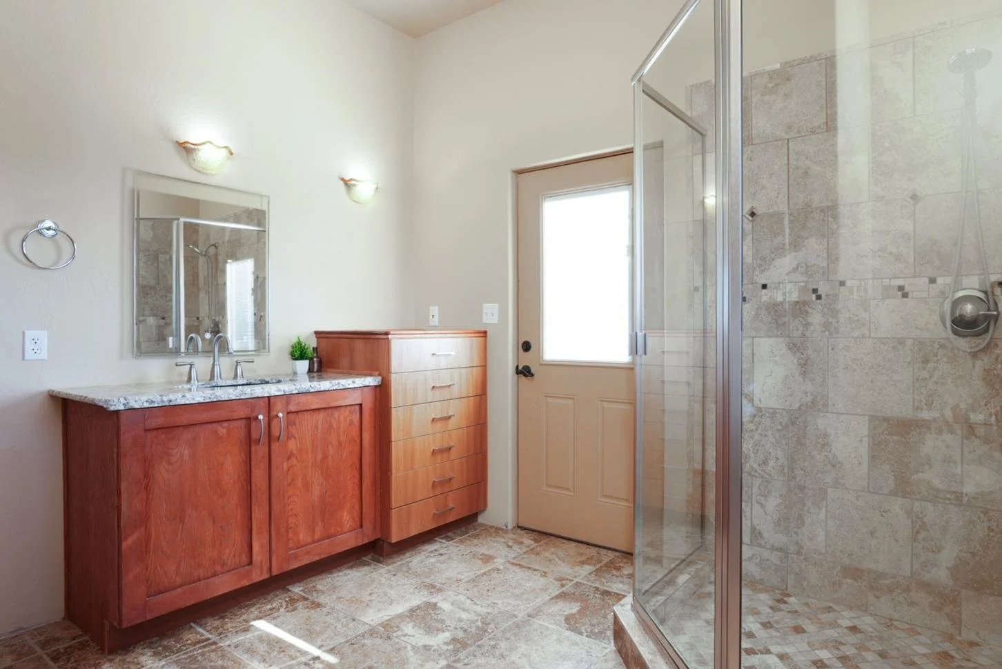 Bathroom with a wooden vanity, granite countertop, mirror, wall-mounted lights, small potted plant, wooden storage cabinet, door with window, and a tiled shower with glass door.