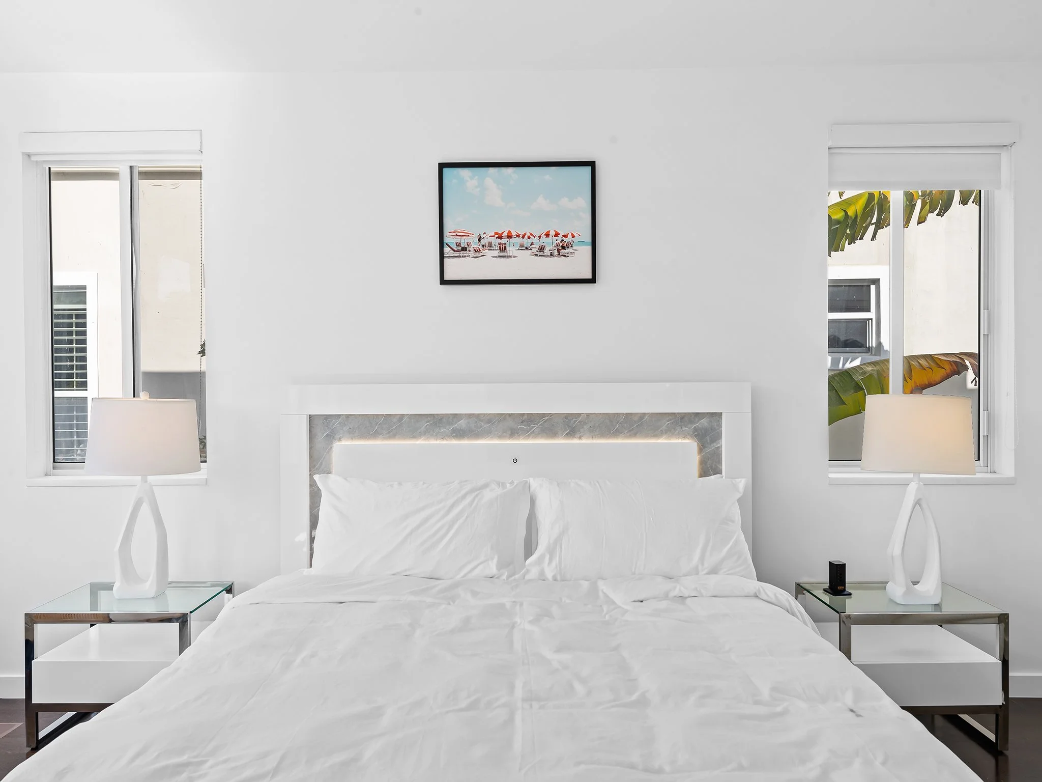 A minimalist bedroom with a white bed, a framed beach scene picture above, two nightstands with modern lamps, and windows with blinds showing a glimpse of trees and buildings outside.