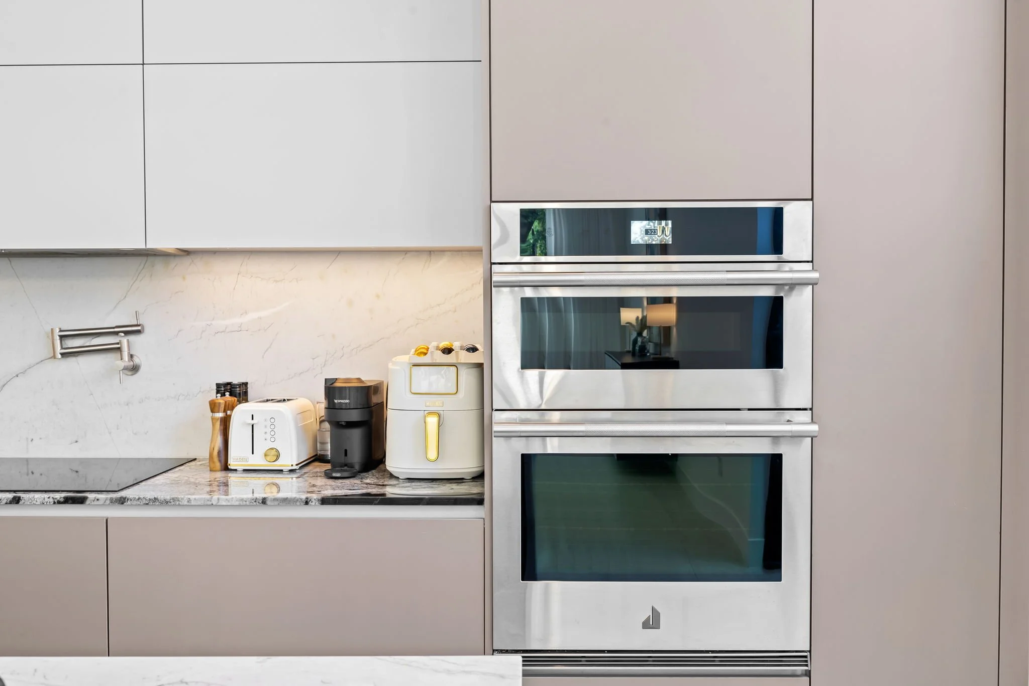 Modern kitchen with built-in oven, microwave, and toaster on marble countertop, beige cabinets, and stainless steel fixtures.