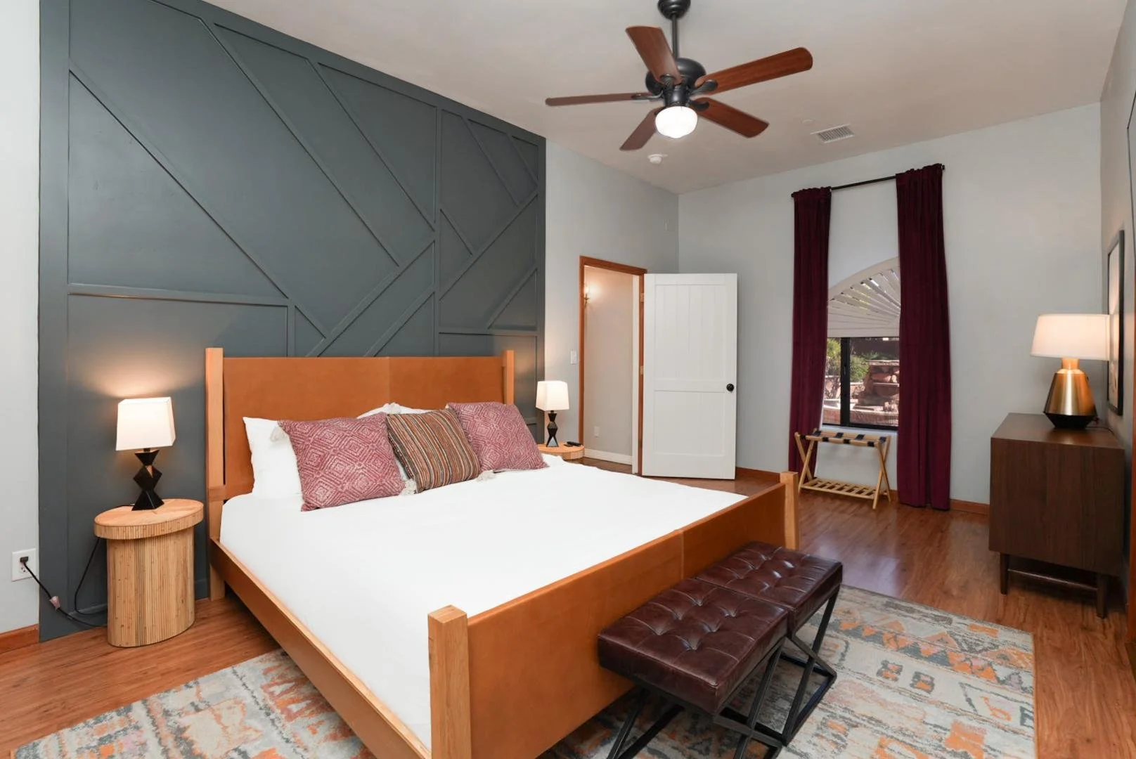 Modern bedroom with a wooden bed frame, quilted headboard, and multiple pillows. There is a dark wood bench at the foot of the bed, and side tables with lamps. The room has hardwood floors, patterned area rug, a window with dark red curtains, and a c