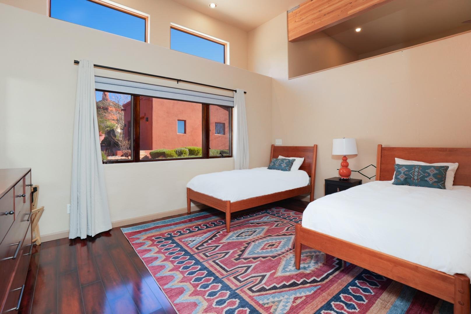 A bedroom with two wooden beds, a nightstand with a white lamp, a colorful geometric patterned rug, and a large window with white curtains showing an outdoor view of a red building and greenery.