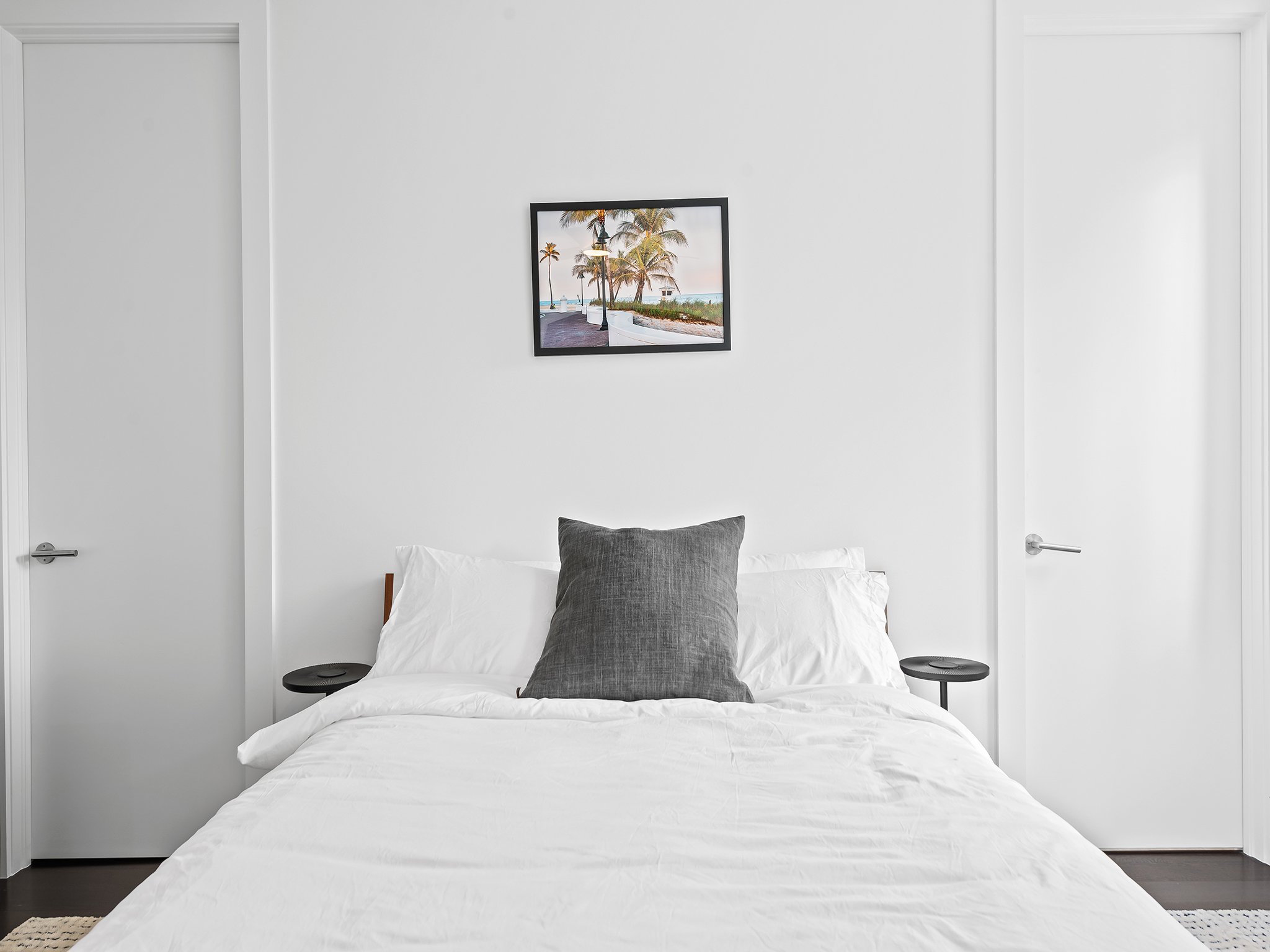 A minimalist white bedroom with a bed, black side tables, and a framed picture of a beach scene with palm trees on the wall.