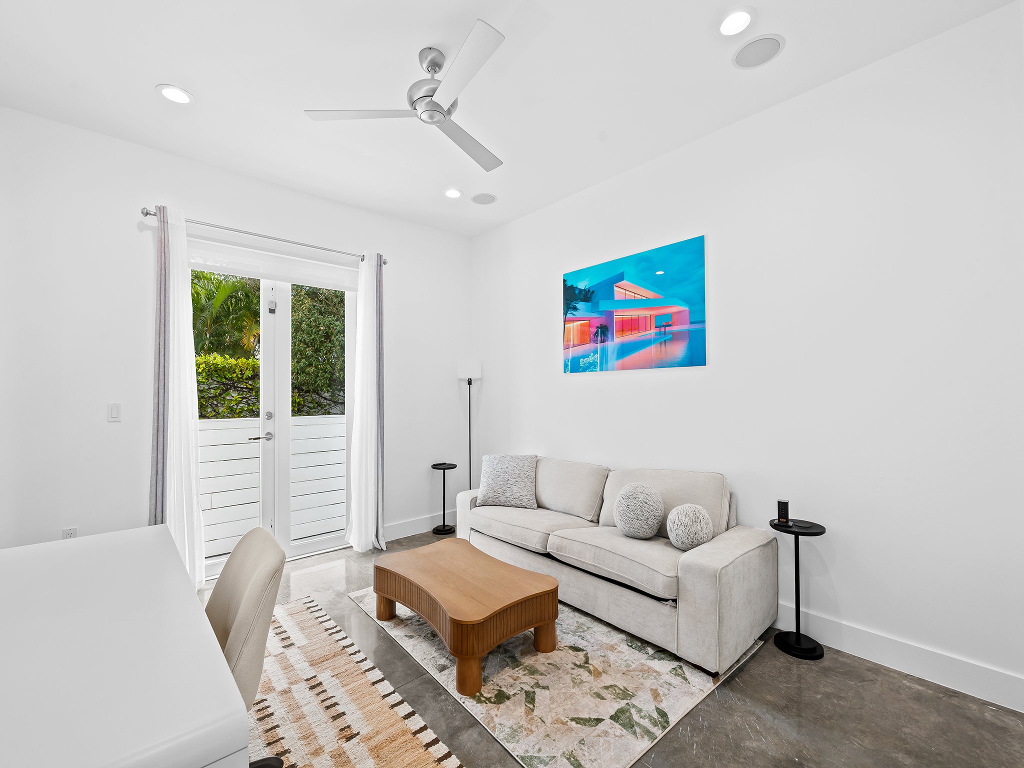 A modern living room with a white sofa, a wooden coffee table, and a colorful abstract artwork on the wall. There is a sliding glass door with white curtains, leading outside to greenery. The ceiling has a silver fan and recessed lighting. A black st