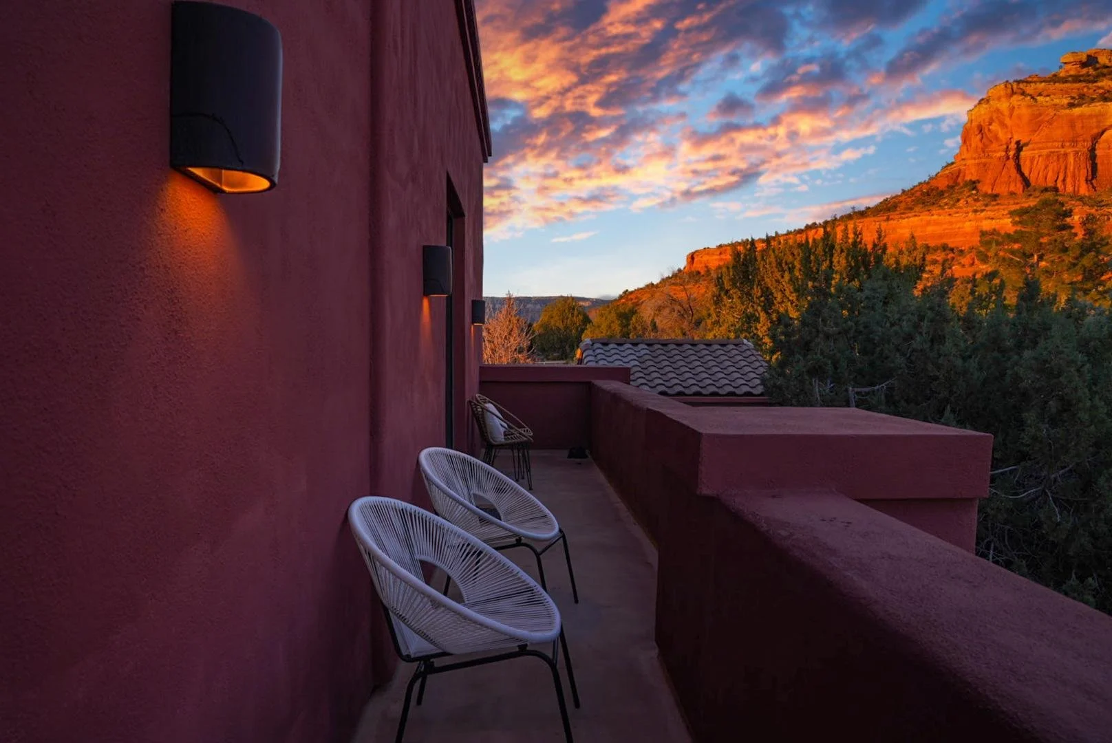 Balcony with three white chairs overlooking a scenic landscape of trees and orange rock formations during sunset.