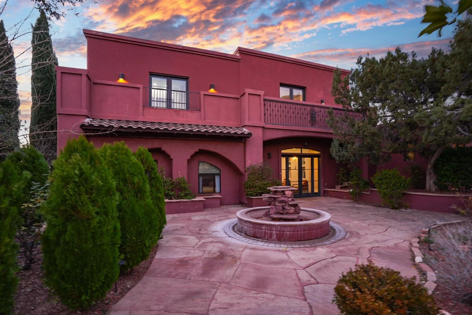 A two-story house with a pink exterior, a balcony, and a fountain in the front yard. The sky has colorful clouds at sunset.
