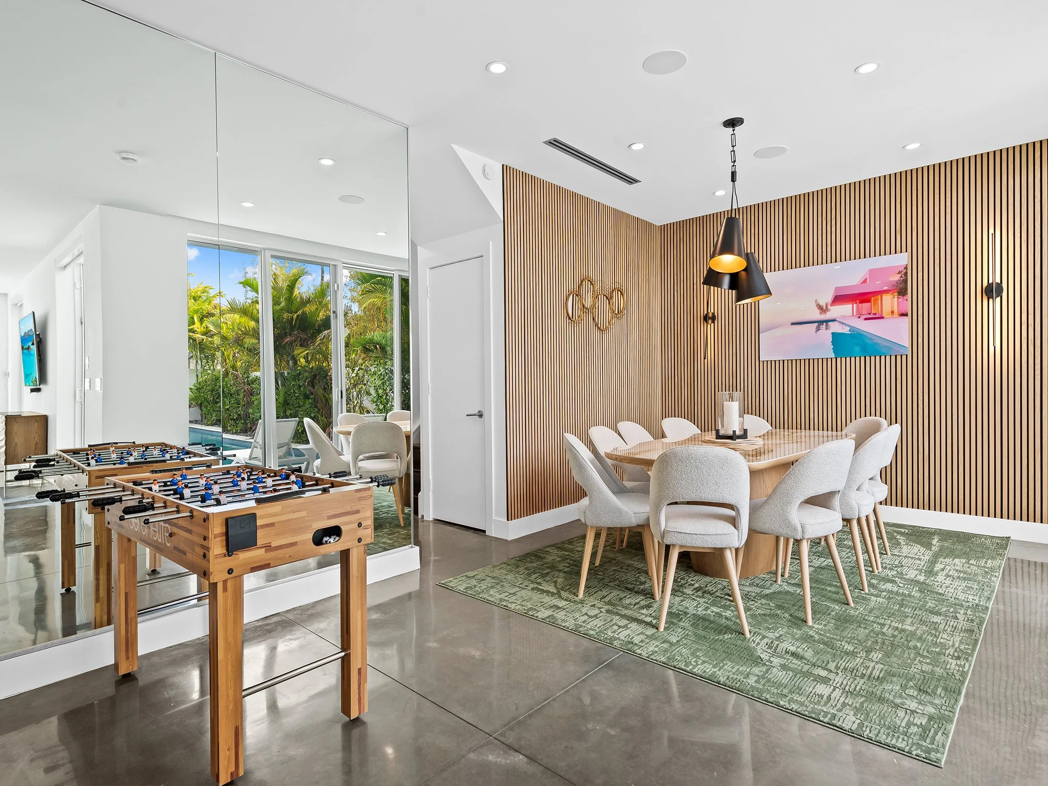 Modern living room with white walls and ceiling, wooden slat accent wall, round dining table with cream upholstered chairs, black pendant lights, large flat-screen TV on the wall, glass doors leading to outdoor greenery, foosball table, and green pat