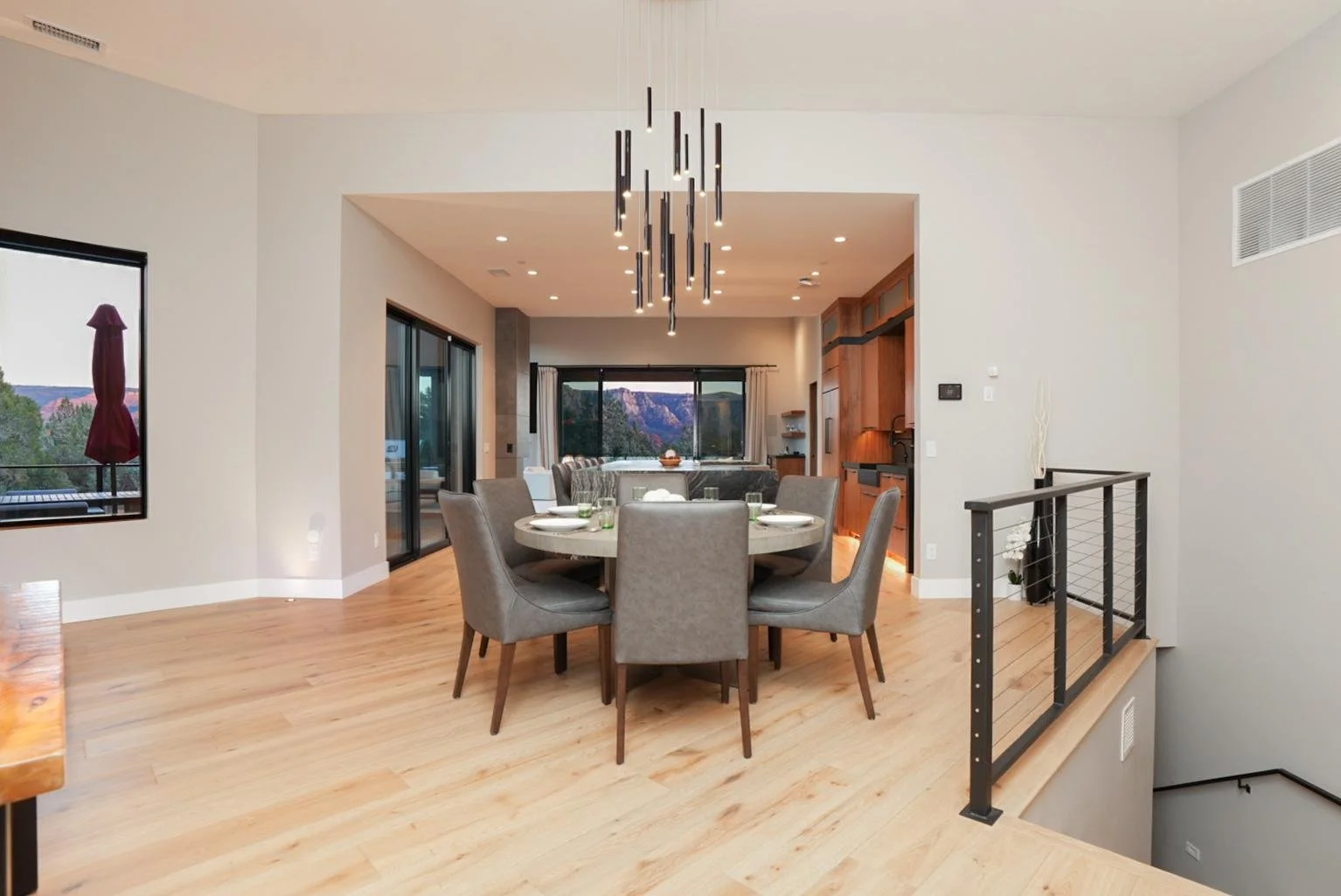 Dining area with a round table and six gray upholstered chairs, wooden flooring, modern chandelier, large sliding glass doors, and views of mountains outside.