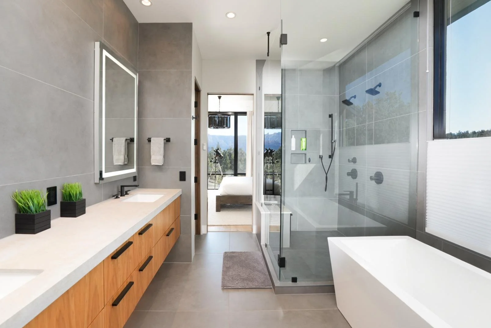 Modern bathroom with a large vanity, mirror, potted plants, and a glass-enclosed shower with multiple showerheads, a view of a bedroom and mountains outside through sliding glass doors.