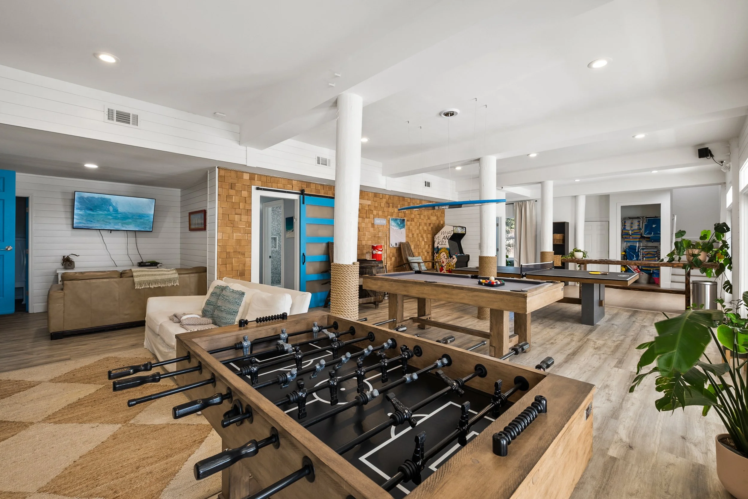 Interior of a recreational room with foosball table, pool table, ping pong table, arcade machine, and seating area with couches and a TV, decorated in a modern style with white walls, wooden accents, and large windows.