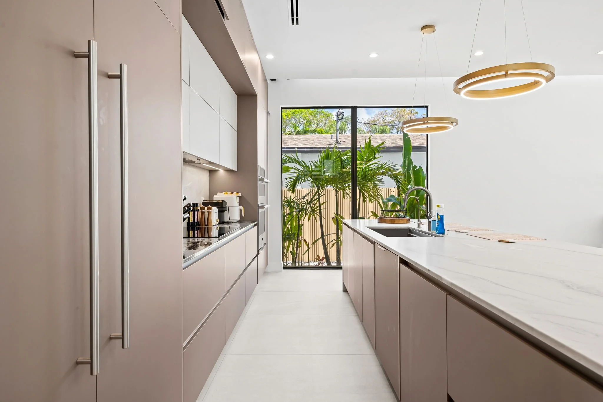 Modern kitchen with white cabinets and marble countertops, large window with view of plants and trees outside, ceiling with circular gold pendant lights.