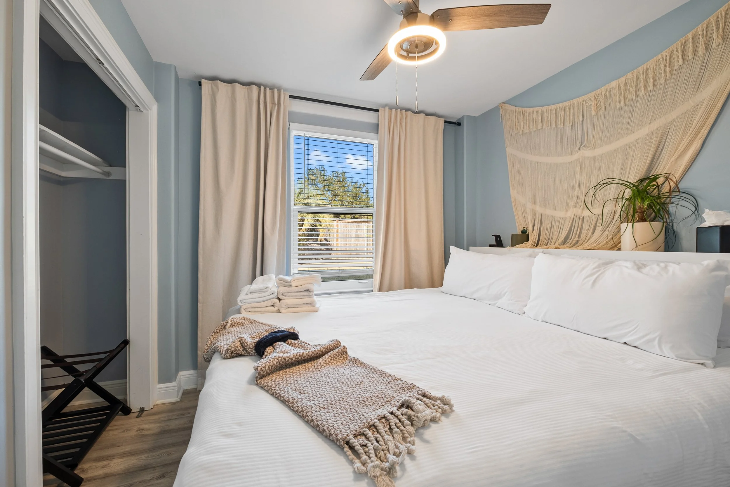 Clean bedroom with a bed with white linens, a window with beige curtains, a ceiling fan, a potted plant, and folded towels.