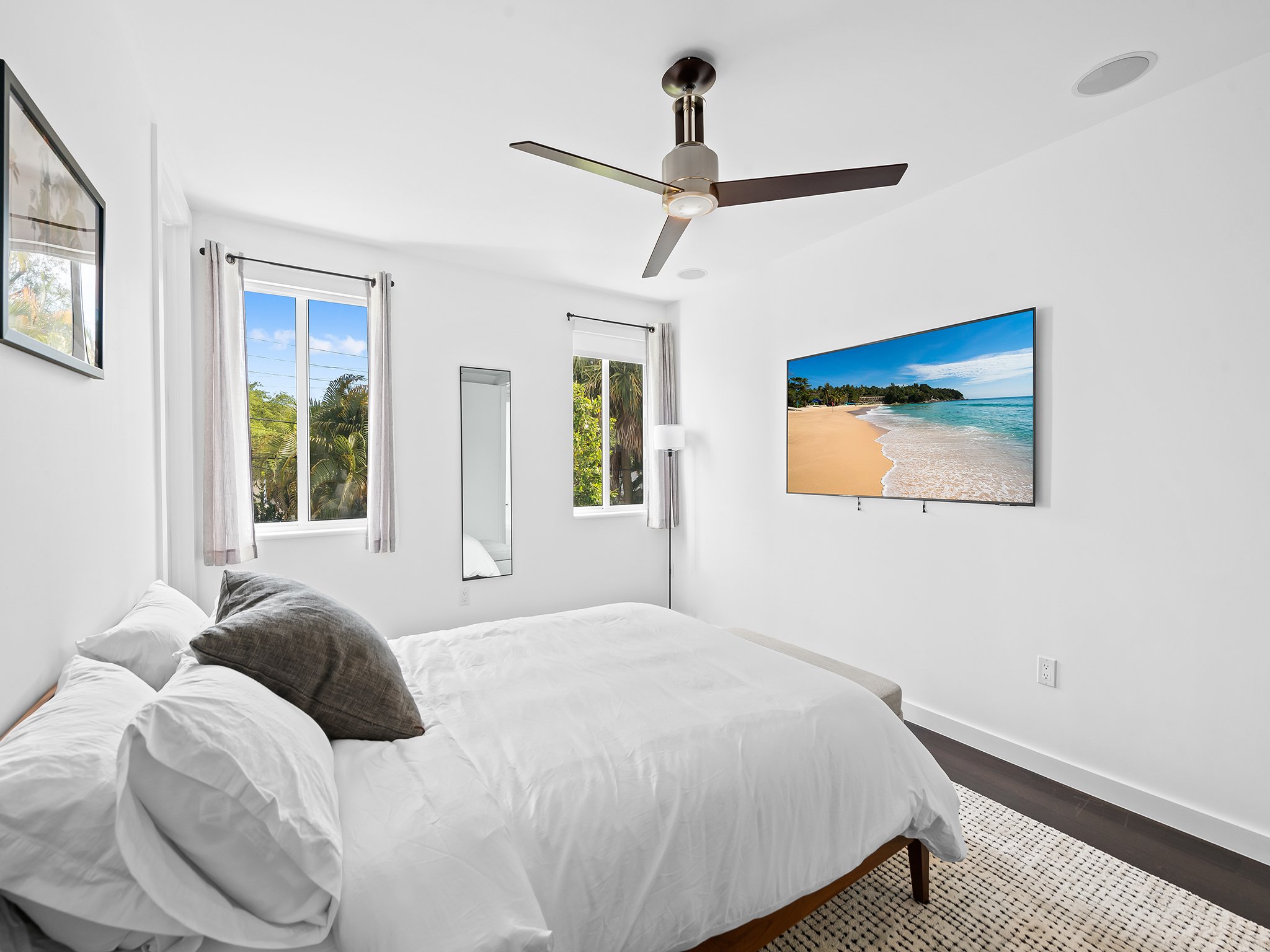 A bedroom with white walls and bedsheets, featuring a wall-mounted TV showing a beach scene, two windows with white curtains, and a ceiling fan.