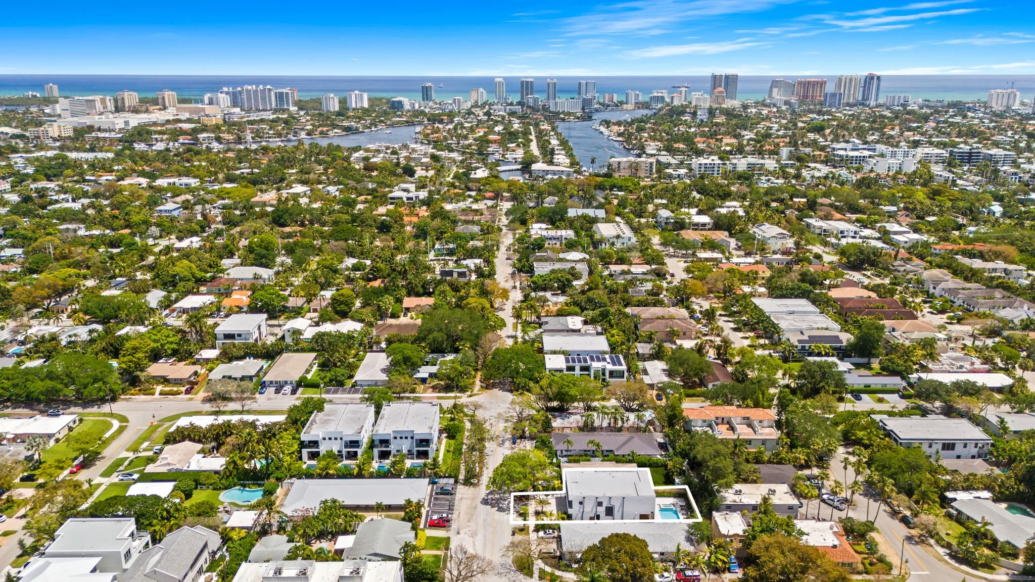 Aerial view of a city with a mix of residential houses, tall buildings, water canals, and the ocean in the background, under a partly cloudy sky.