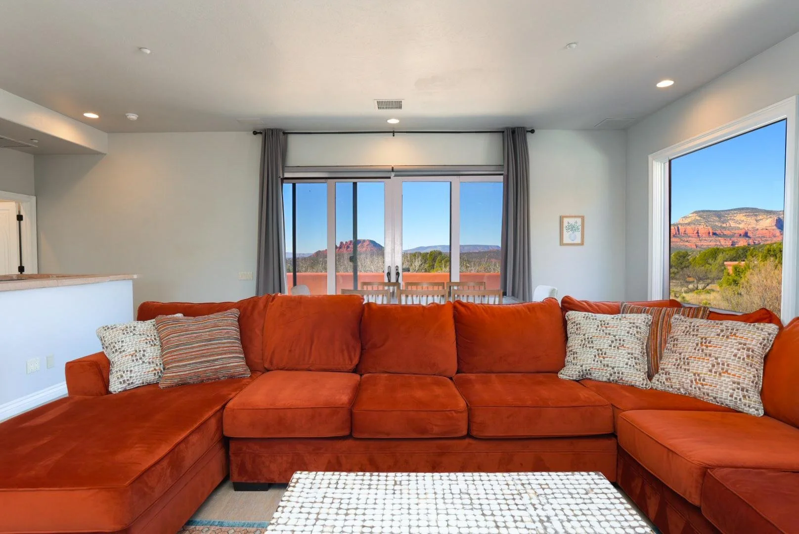 Living room with an orange sectional sofa, patterned throw pillows, large windows, outdoor view of mountains and clear sky, dining table, and curtains.
