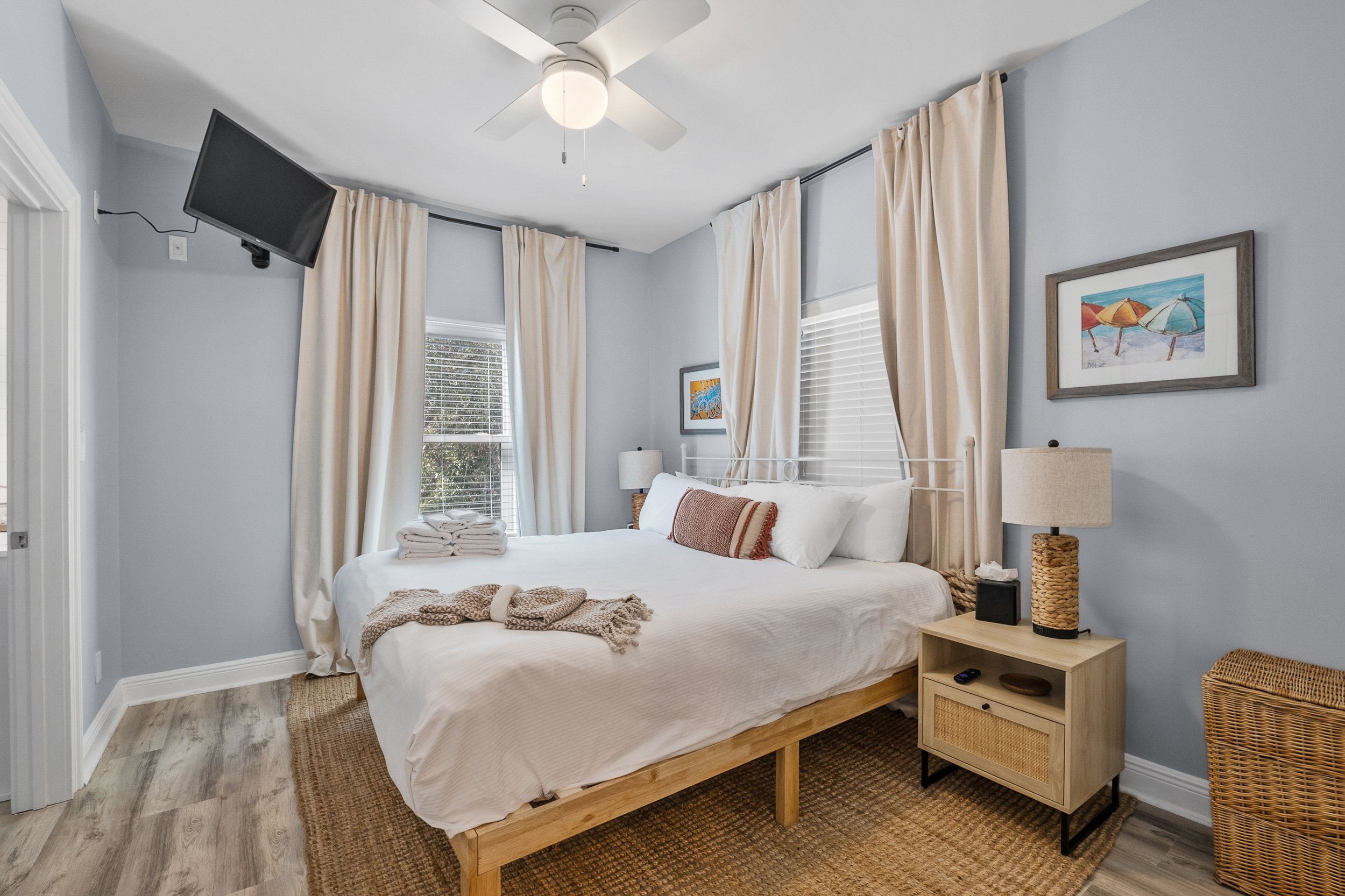 A cozy bedroom with a large bed covered in white linens and pillows, a nightstand with a lamp, and windows with cream curtains. A small flat-screen TV is mounted in the upper corner. Decor includes framed beach-themed artwork.
