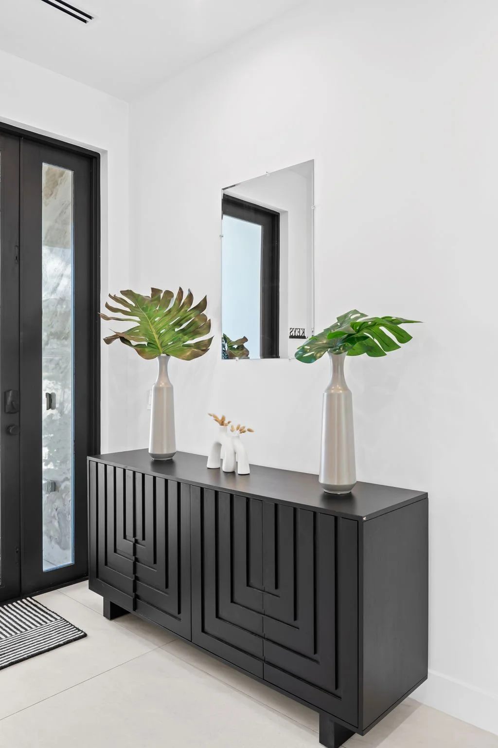 Modern black sideboard with two large vases holding green monstera leaves and two small ceramic decorative items, against a white wall with a rectangular mirror and a window.