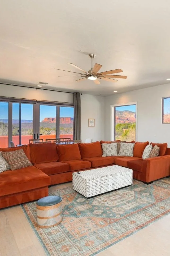 Living room with a large orange sectional sofa, patterned throw pillows, a white woven trunk coffee table, and a patterned area rug, with large windows showing a scenic view of red rock formations.
