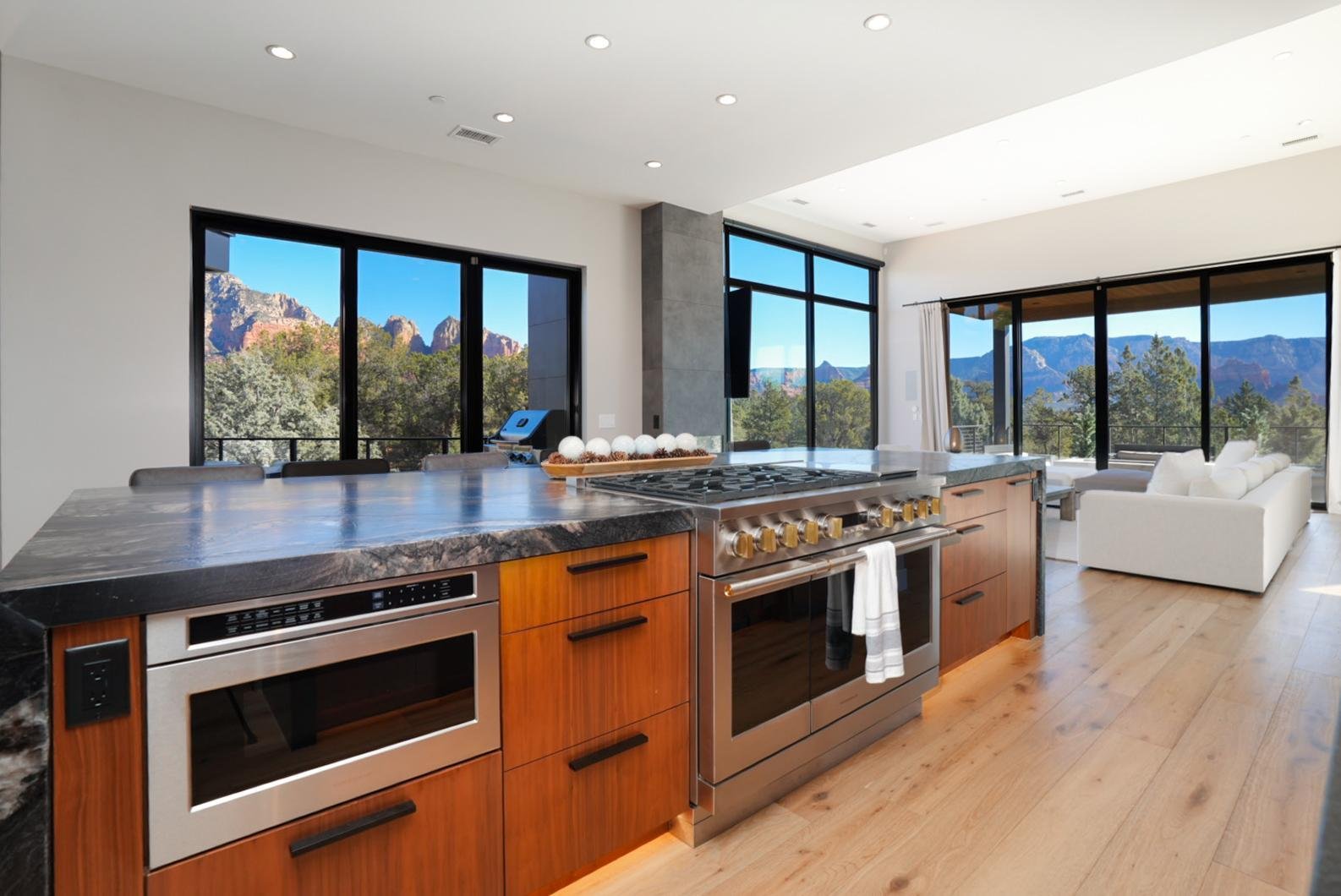 Modern kitchen with a view of mountains and trees through large windows, featuring wooden cabinets, a granite countertop, and stainless steel appliances.