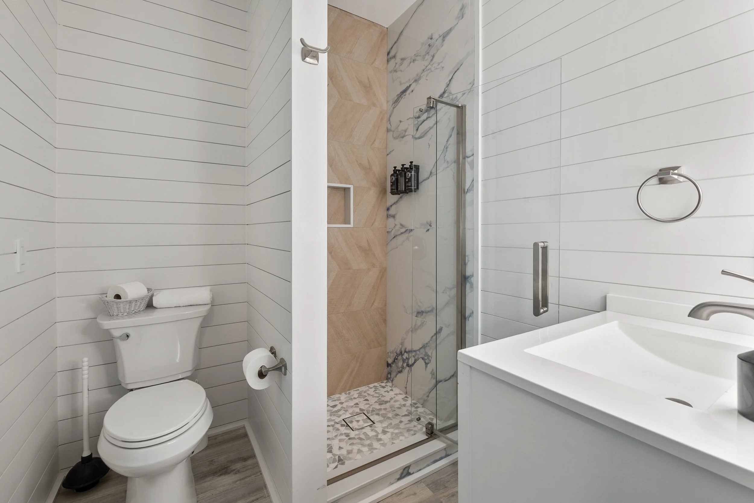 A modern bathroom with white horizontal shiplap walls, a shower with marble and wood-look tiles, a toilet with a basket of toilet paper on top, and a white vanity with a sink. The shower has a glass door and black soap dispensers inside.