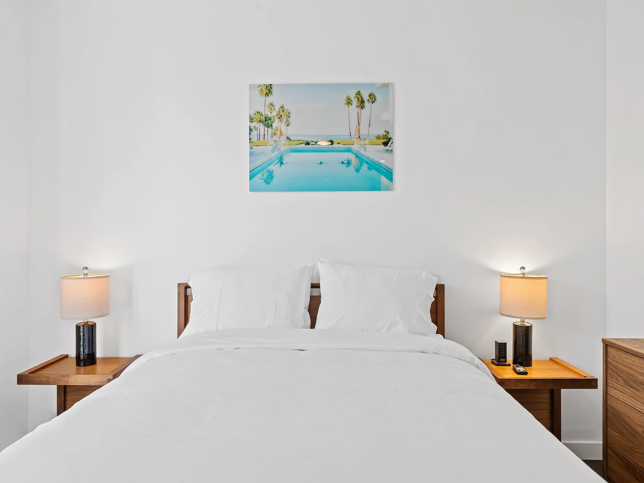 Bedroom with white bed, wooden nightstands, two lamps, and a framed coastal landscape painting above the bed.