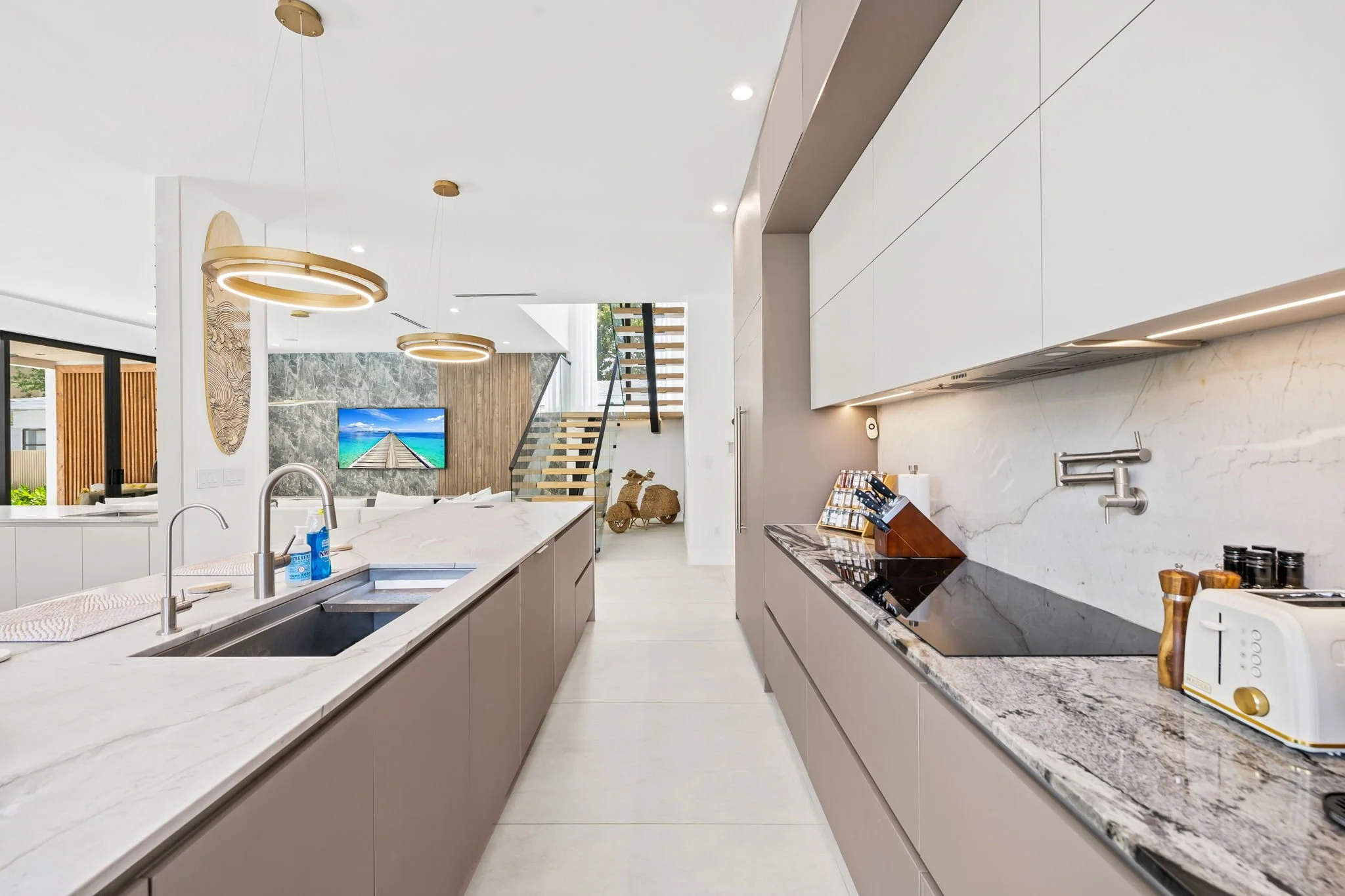 Modern kitchen with white countertops, beige cabinets, and a marble backsplash. There is a double sink, a black cooktop, and a toaster on the counter. Bright lighting and large windows provide natural light.