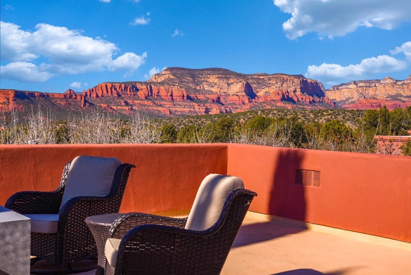 Outdoor patio with wicker chairs and cushions overlooking red rock mountains under blue sky with clouds.