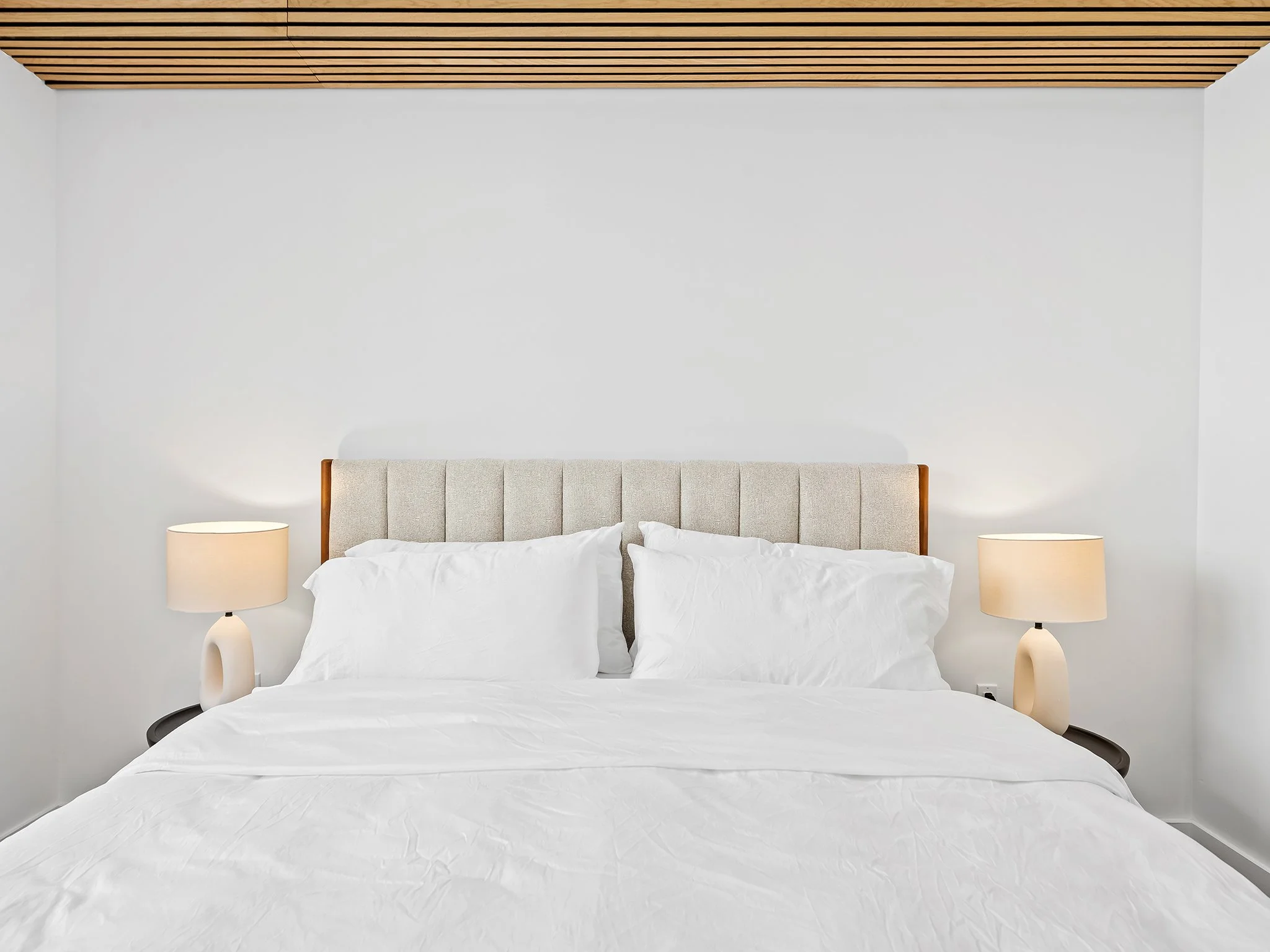 A modern bedroom with a beige upholstered headboard, white bedding, two beige bedside lamps, and a wooden slat ceiling.