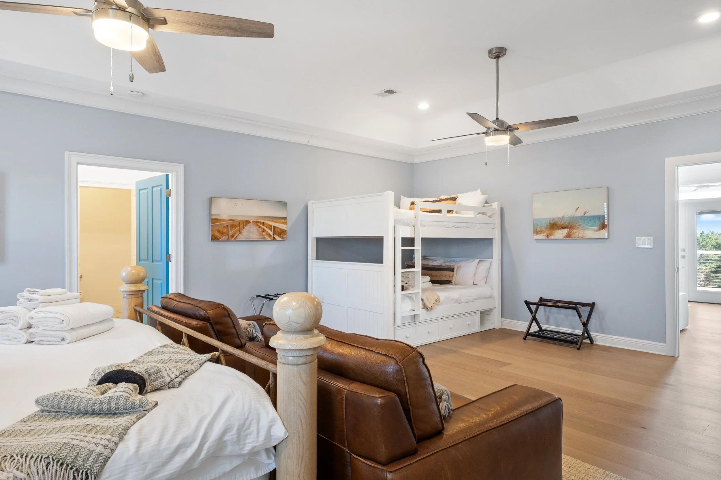 A bedroom with light blue walls and hardwood floors, featuring a bunk bed, a leather sofa, a bed with white linens, ceiling fans, and wall art.
