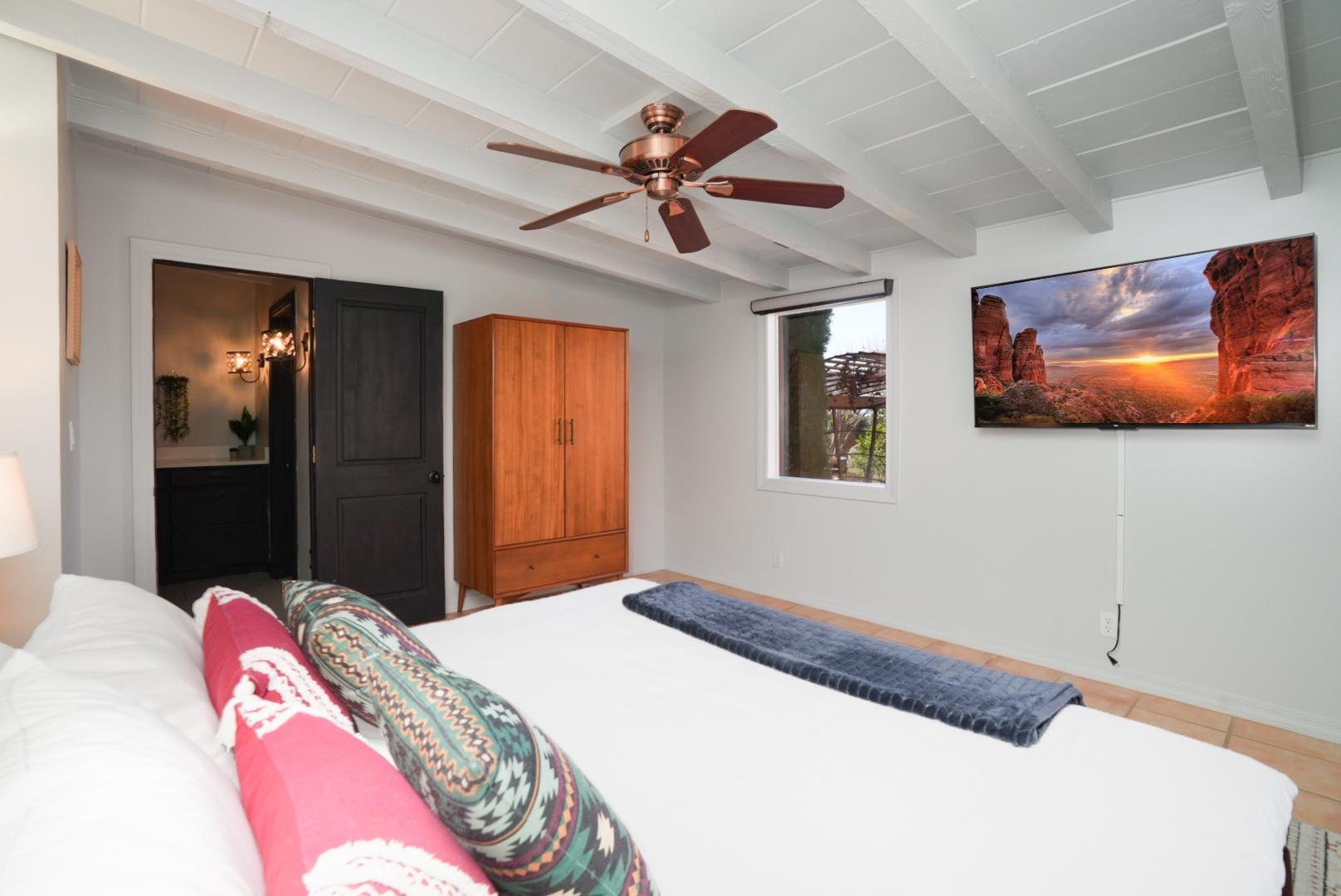 A bedroom with a white ceiling and light-colored walls. There is a ceiling fan with wooden blades. A flat-screen TV is mounted on the wall, showing a scenic landscape of cliffs and a sunset. A window with a view outside. Part of a bed with white bedding and colorful pillows is visible in the foreground.