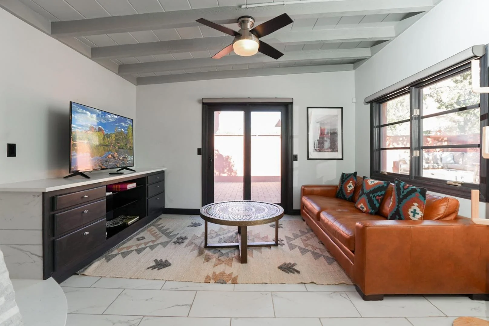 Living room with a leather sofa, patterned pillows, a wooden coffee table, a flat-screen TV on a black and white console, a framed picture on the wall, large windows, a sliding glass door leading outside, a ceiling fan, and a rug on the tiled floor.