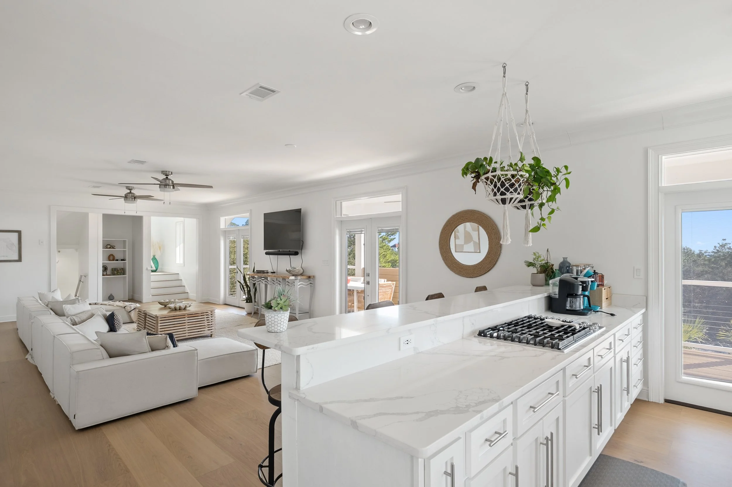 Open concept living room with white sofa, TV, and a white kitchen with a marble countertop, hanging plants, and patio door with a view outside.