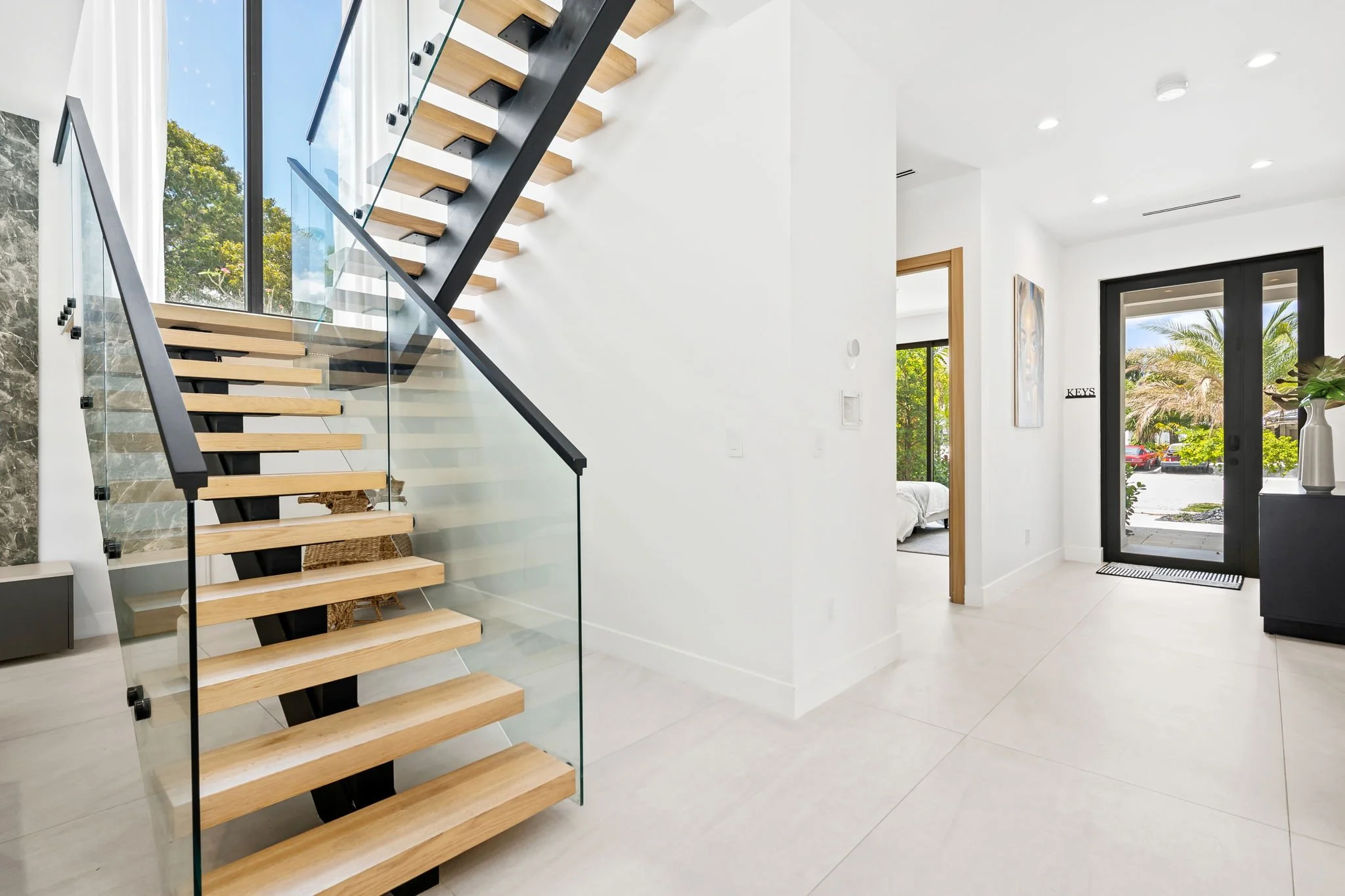 Modern staircase with wooden steps, glass railing, and metal support in a bright, open interior with large windows, white walls, and minimal decor.