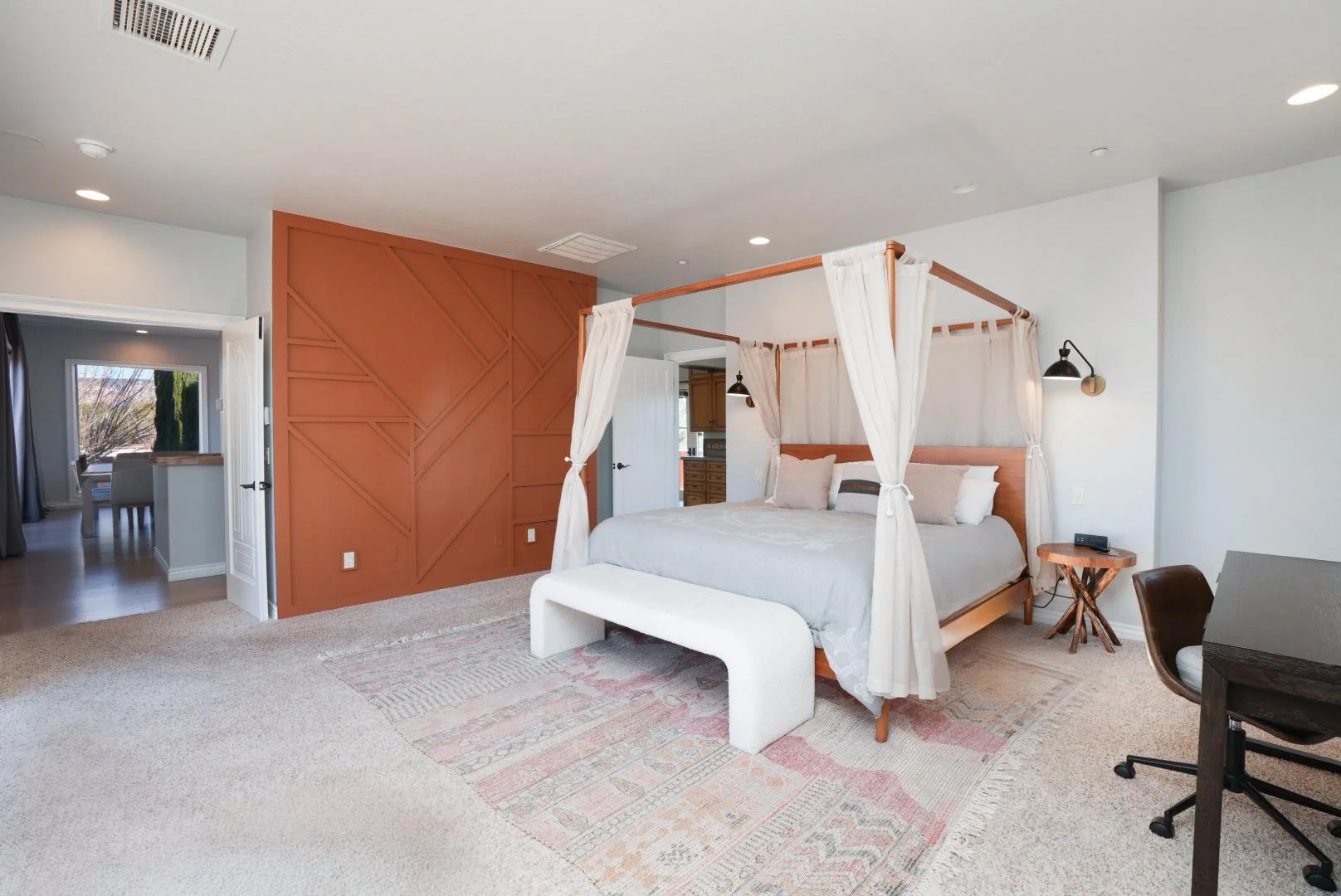 Bedroom featuring a four-poster bed with white curtains, a wooden nightstand, and a patterned area rug, with an open door leading to a living space.