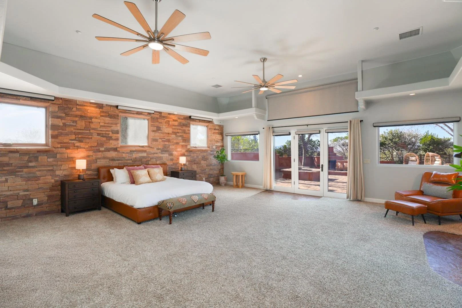 Bright bedroom with a brick accent wall, a bed with tan and pink pillows, black nightstands with lamps, a bench at the foot of the bed, sliding glass doors leading to a patio, and a tan armchair with an ottoman near a window.