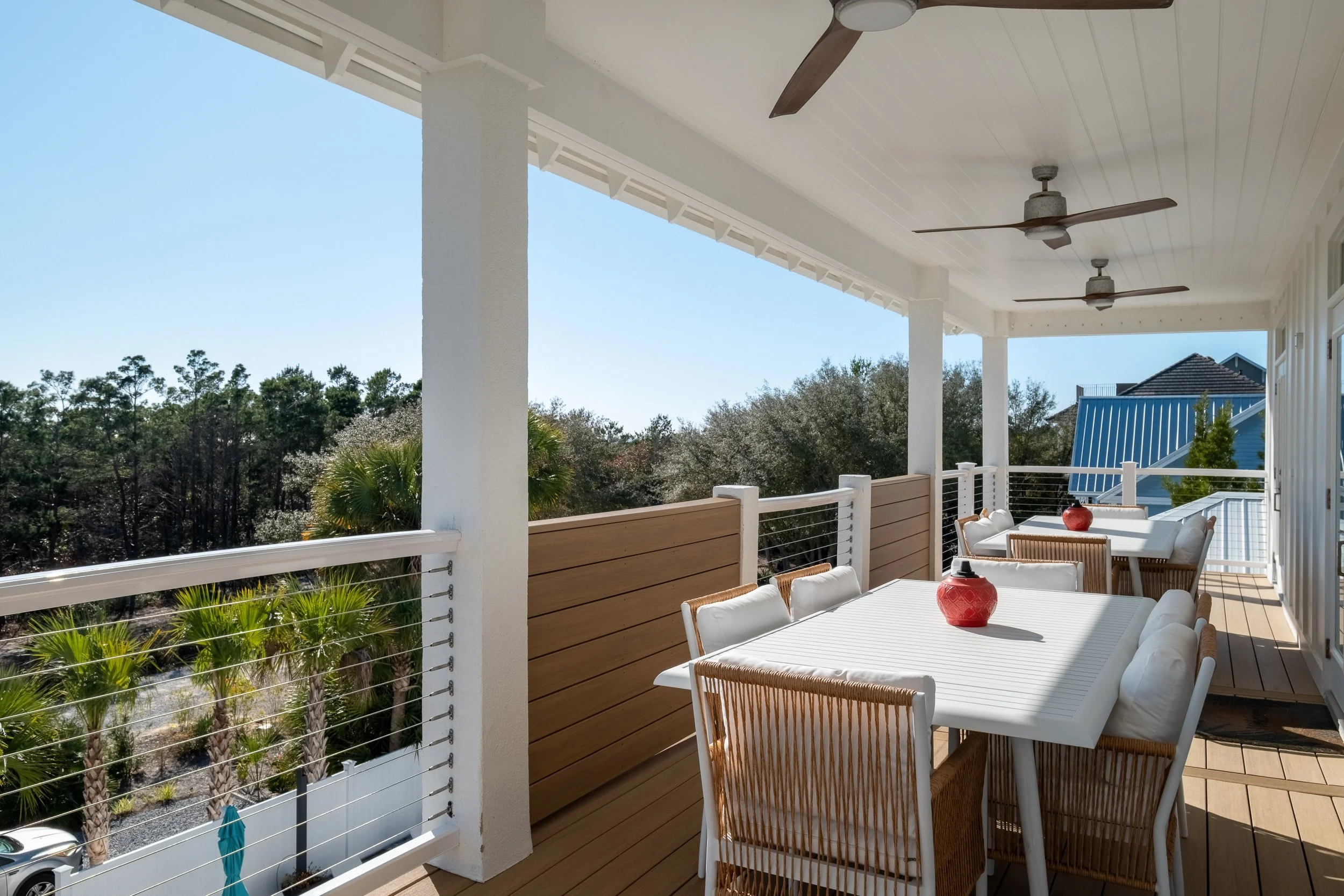 Outdoor balcony with white dining table and chairs, decorated with red vases, overlooking trees and neighborhood rooftops, featuring ceiling fans and wooden flooring.
