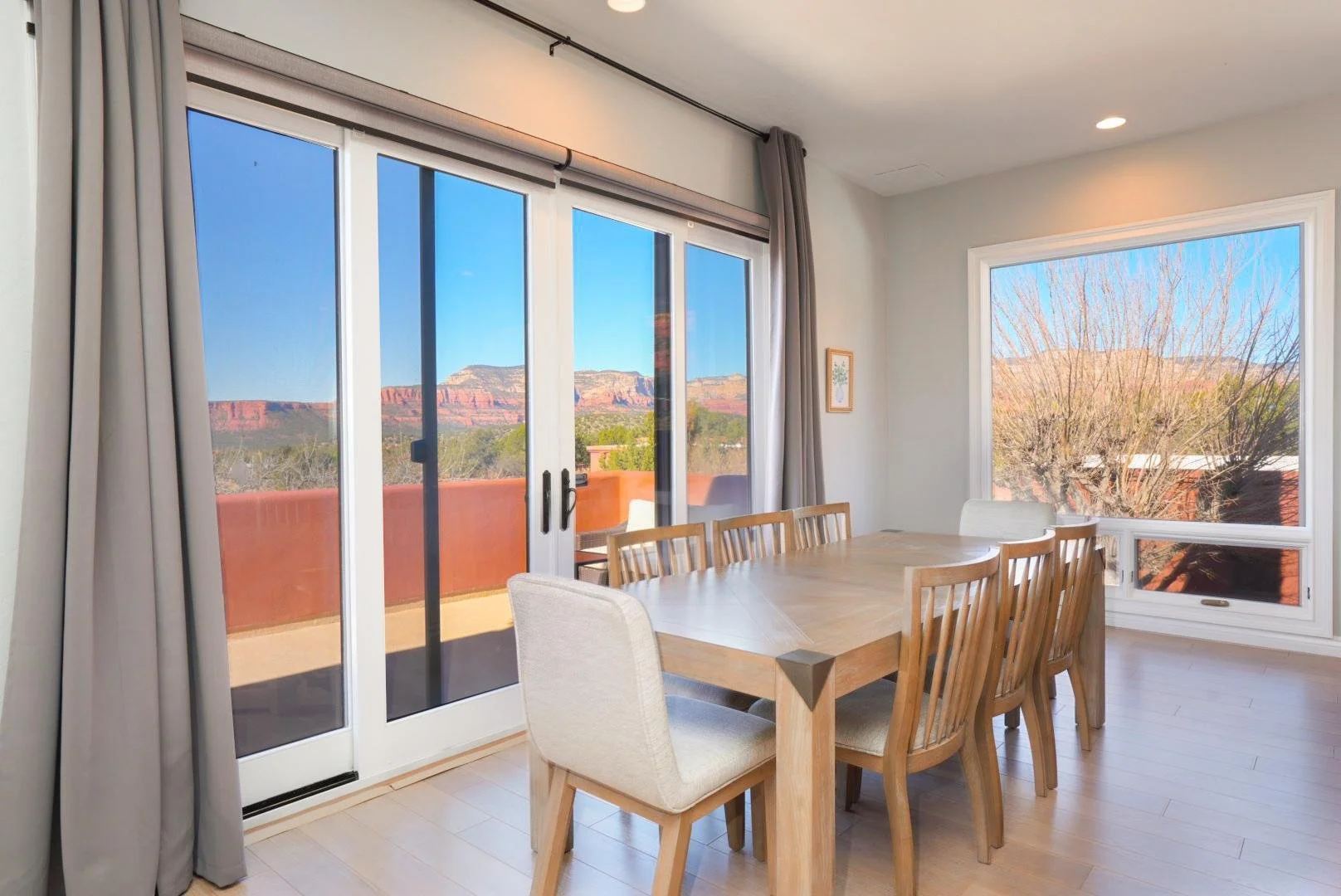 Dining room with large glass sliding doors and a window showing desert mountains in the distance, a wooden dining table with 8 chairs, and a small framed picture on the wall.