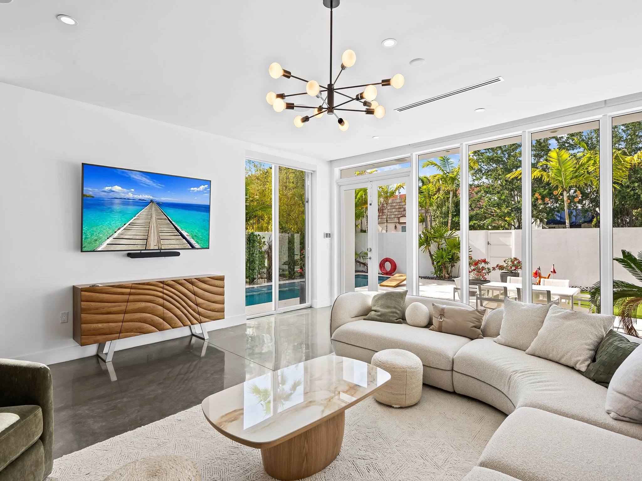 A modern living room with white walls, a cream-colored sectional sofa with pillows, a marble-top coffee table, a wooden sideboard beneath a wall-mounted flat-screen TV showing a pier leading into a blue ocean, a large ceiling light fixture, and large