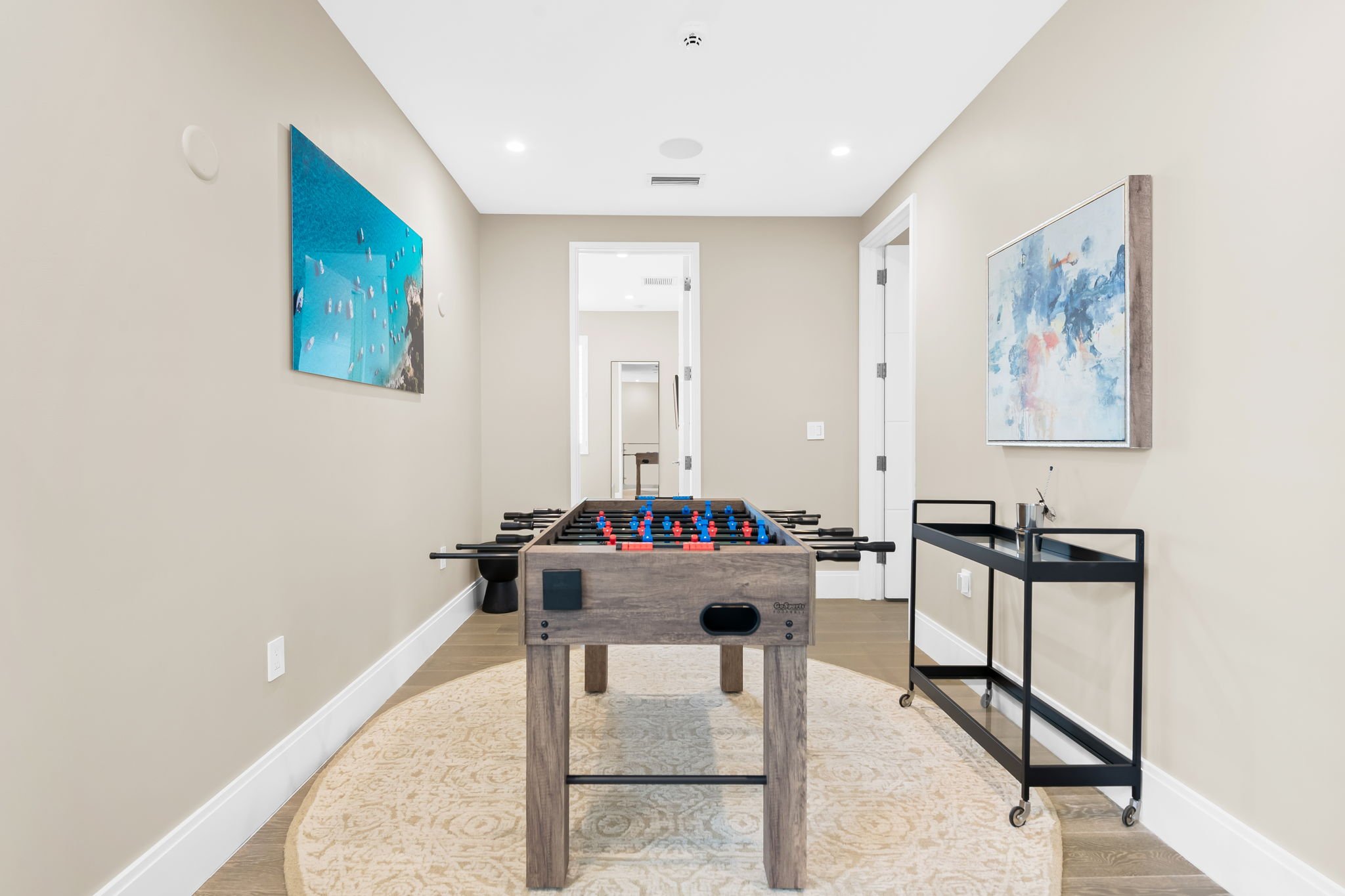 A narrow hallway with beige walls and a white ceiling, decorated with two abstract paintings. It features a foosball table in the center and a black serving cart to the right. The hallway leads to a staircase seen through a door at the end.