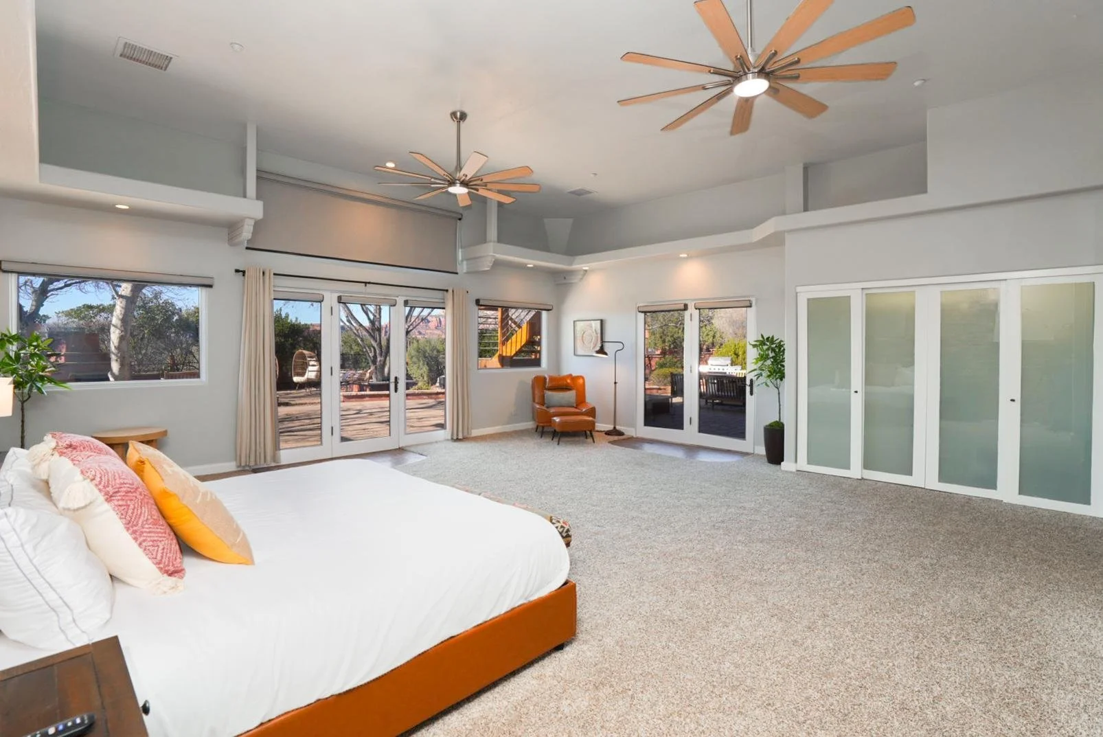Spacious bedroom with large windows, French doors leading outside, a white bed with colorful pillows, a brown armchair, potted plants, and ceiling fans.