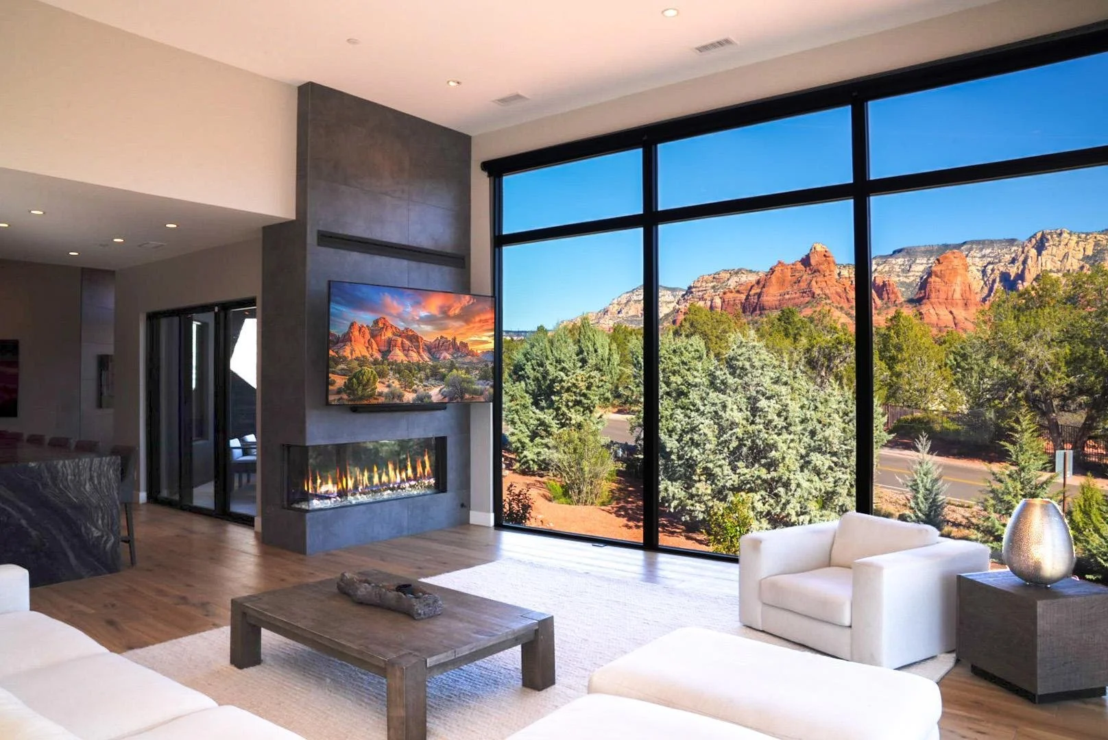 Modern living room with large glass windows showcasing mountains and trees outside, a white armchair, a wooden coffee table with a piece of driftwood, a fireplace, and a wall-mounted television displaying a desert landscape.
