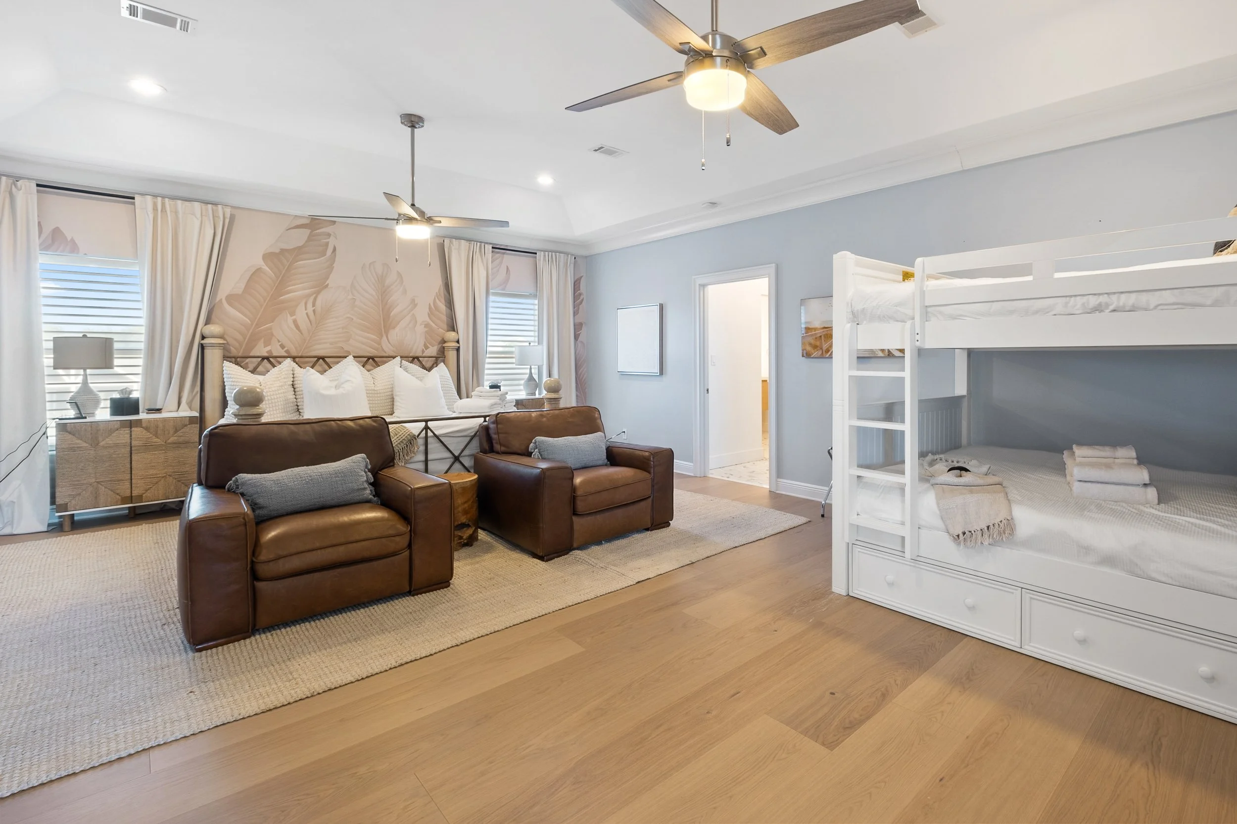 A spacious bedroom with a coastal style, featuring two leather armchairs, a bunk bed with white bedding, a large bed with multiple pillows, and large windows with white curtains, a ceiling fan, and soft lighting.