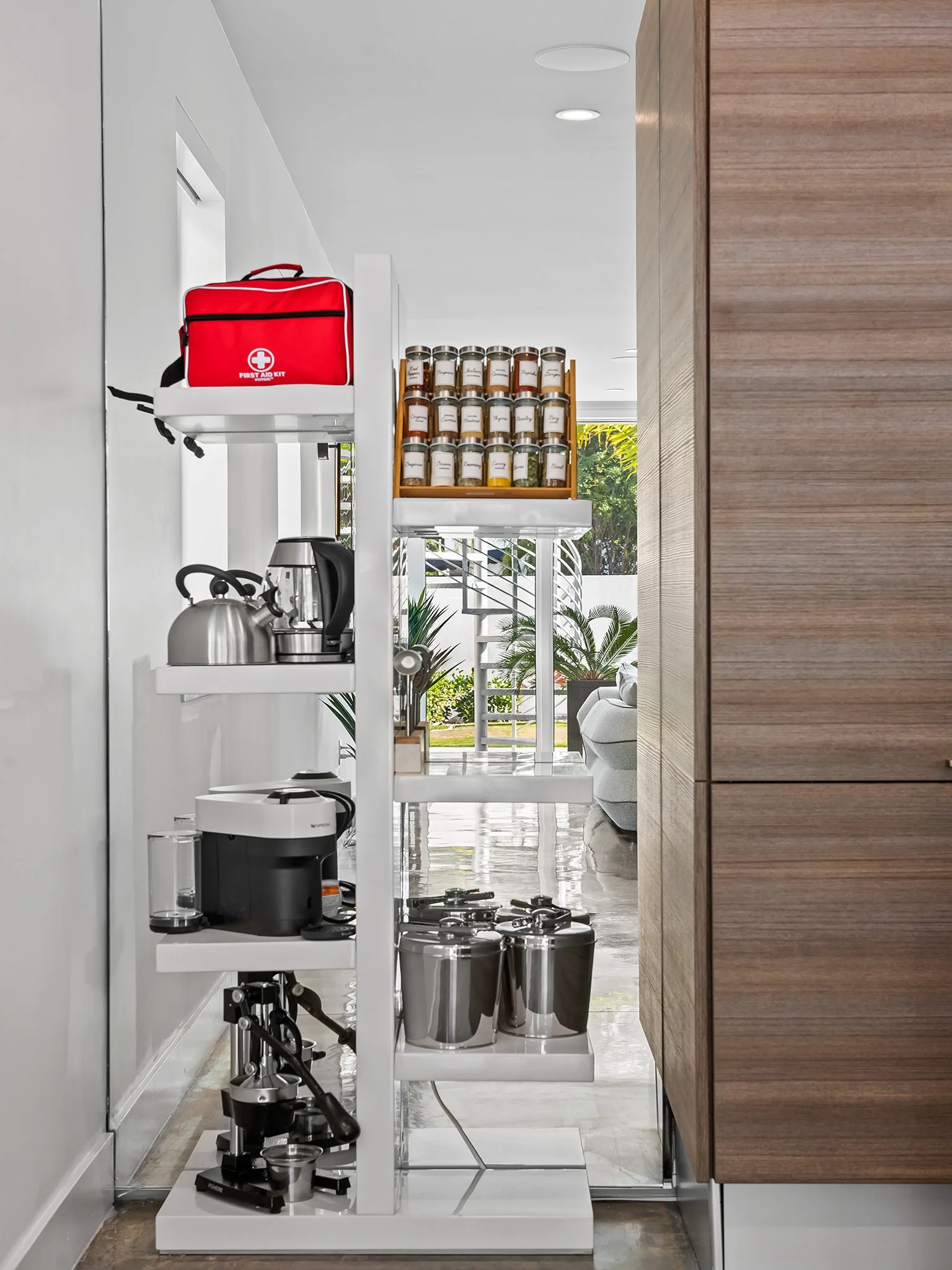 Kitchen shelf with a red first aid kit, multiple spices jars, a silver kettle, a coffee machine, and silver trash cans, with a living room and garden visible in the background.