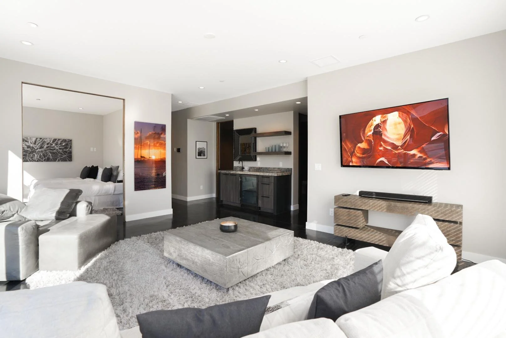 Modern living room with a large white sofa, a matching armchair, and a gray area rug. A framed picture of a sunset over the water hangs on the wall. A flat-screen TV displays a canyon scene. A small kitchen area with bar shelves and countertop is vis