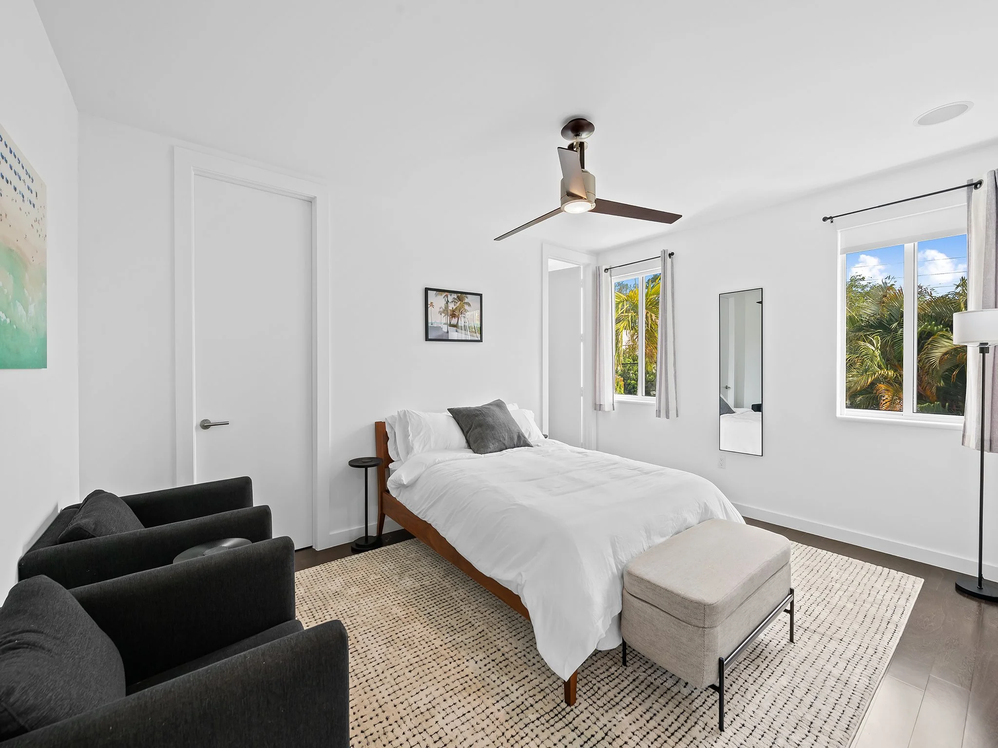 A bedroom with white walls and ceiling, a ceiling fan, two windows with white curtains showing greenery outside, a framed picture on the wall, a bed with white bedding and gray pillows, a small black side table, a black armchair, an ottoman with a be